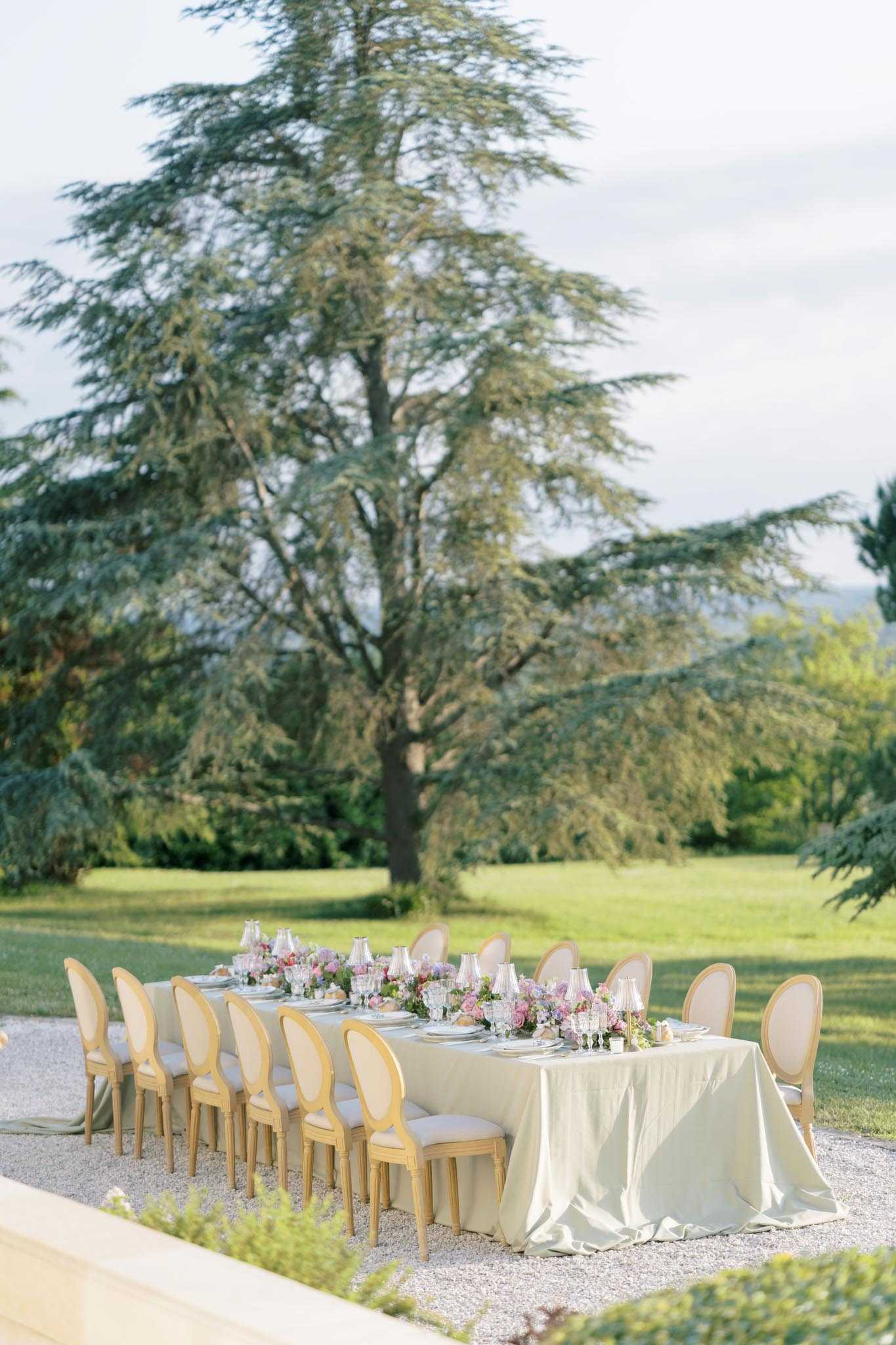 An outdoor wedding reception tablescape set on a gravel terrace surrounded by manicured gardens. The long rectangular table is dressed with an ivory linen tablecloth that pools slightly on the ground, and is lined on both sides with gold-framed Louis XVI oval-back chairs with cream upholstered seats and backs, seating approximately 12 guests. The centerpiece runs the length of the table and features clusters of pink and mauve roses, lavender, and soft purple blooms with greenery, accompanied by glassware including tinted pink goblets and crystal stemware, along with white ceramic place settings. The overall decor palette is soft ivory, gold, and pastel pink and purple, reflecting a classic French romantic styling. Wide shot taken from a slight elevation, with no people present.