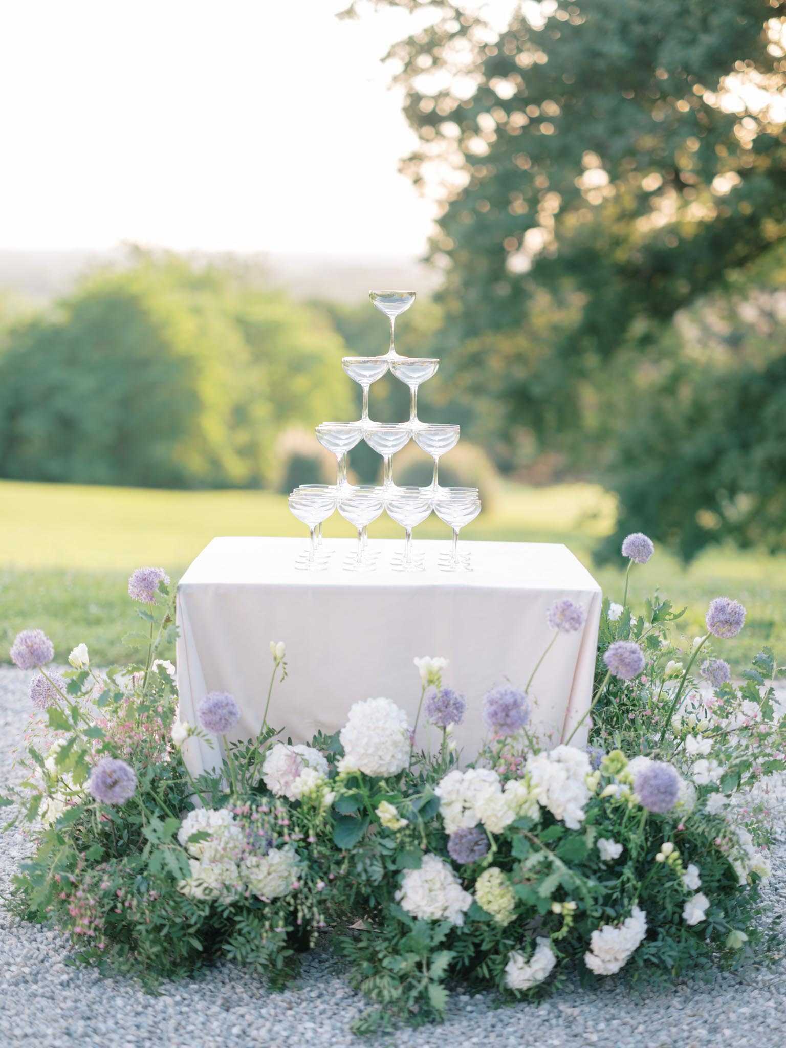A champagne coupe tower is arranged on a small square table covered with a blush pink linen, set outdoors on a gravel surface during what appears to be a cocktail hour. The tower consists of four tiers of coupe glasses, empty and ready to be filled. The base of the table is surrounded by a lush ground-level floral arrangement featuring white hydrangeas, ivory stock flowers, purple allium spheres, soft pink accents, and abundant green foliage. The composition is a medium wide shot with the background softly blurred, showing open lawn and trees in warm late-afternoon light.