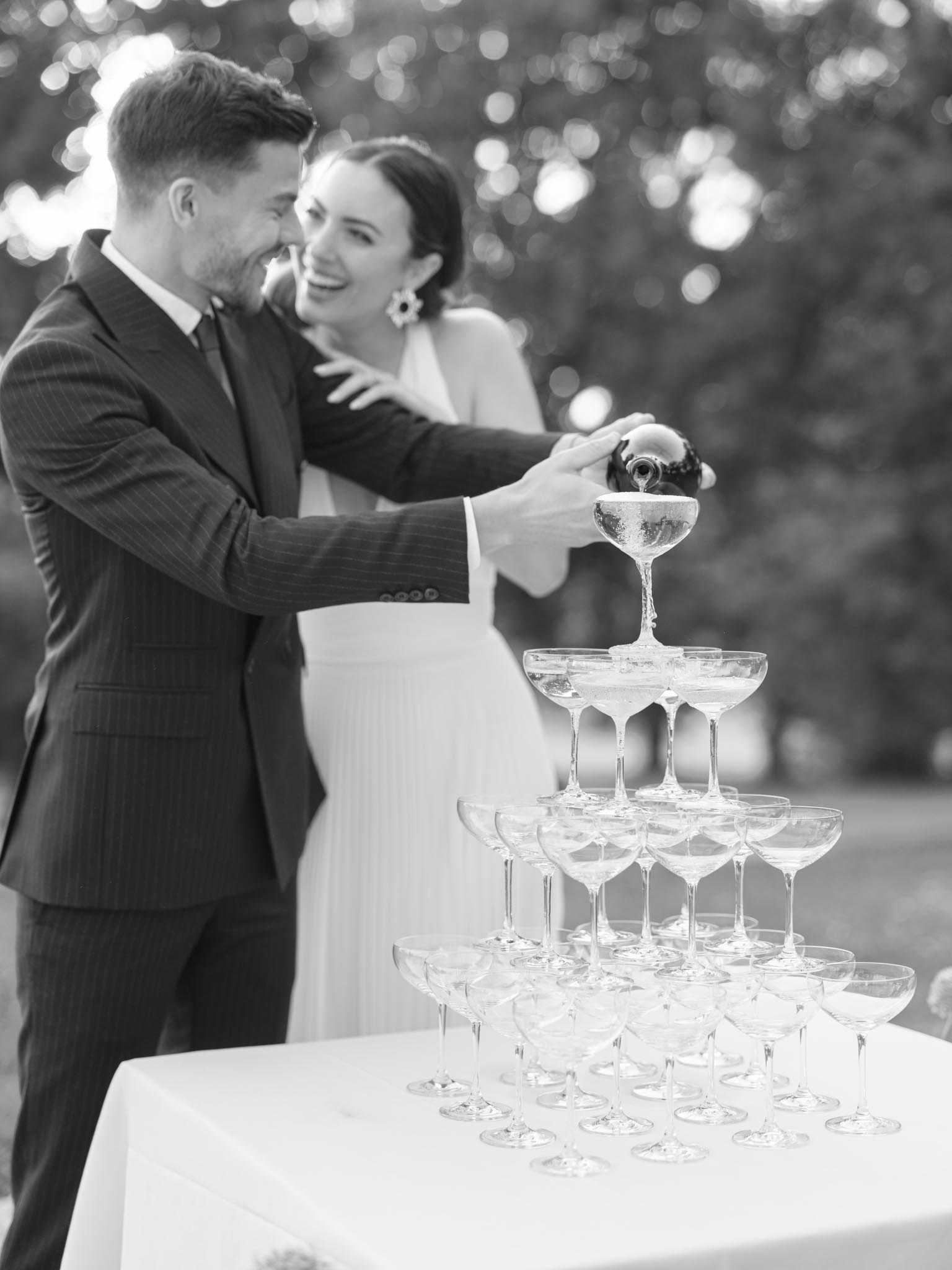 This black-and-white image shows a couple pouring champagne into a coupe glass tower during an outdoor reception. The groom wears a pinstripe suit with a tie, and the bride wears a sleeveless white gown with statement floral earrings, her arm wrapped around his shoulder as both laugh. The pyramid tower consists of approximately five tiers of coupe glasses arranged on a white linen-covered table, with champagne visibly cascading from the top glass downward. The shot is a medium portrait composition with the couple in sharp focus against a softly blurred background of trees and bokeh light spots.