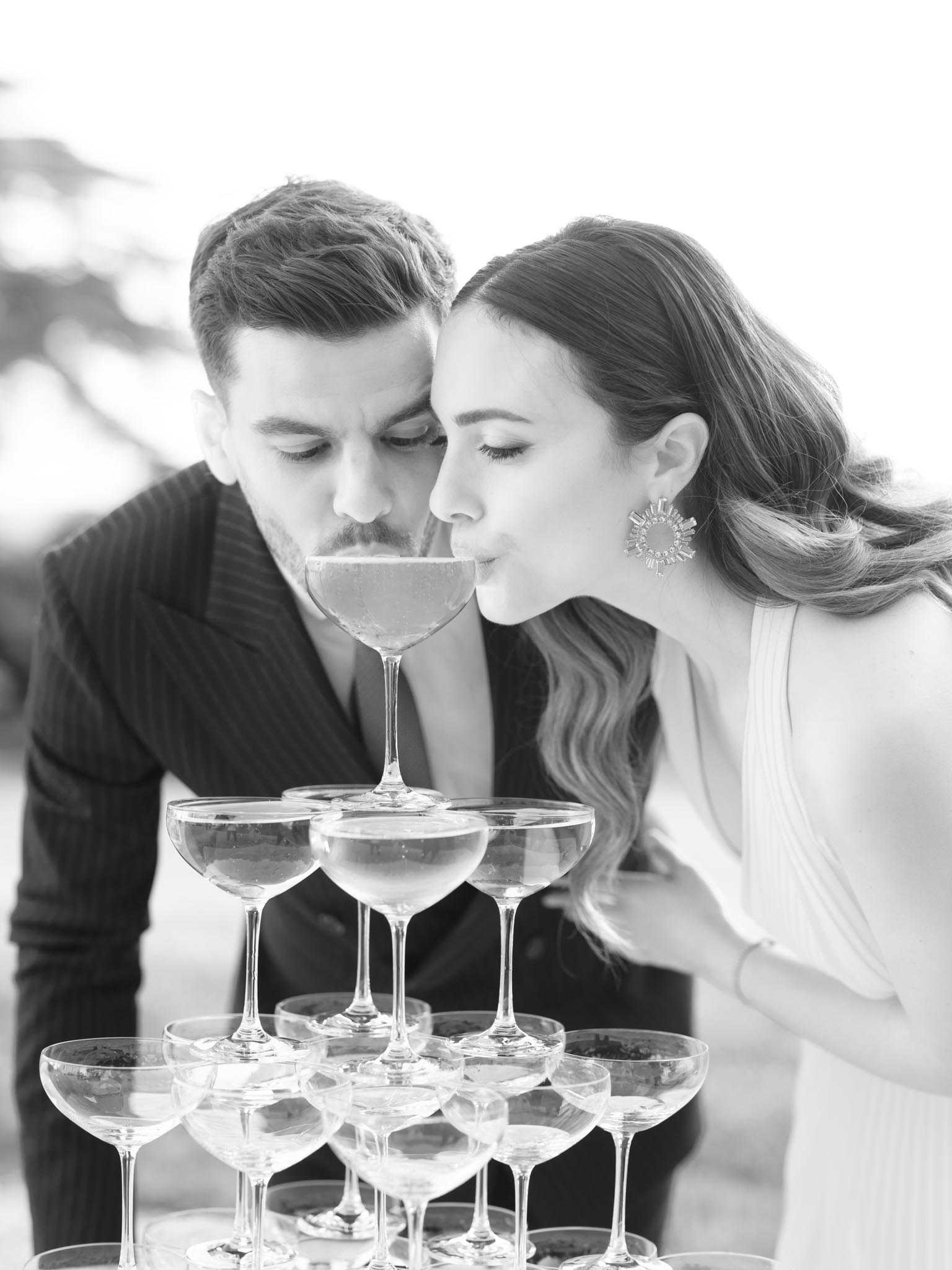 This is a black-and-white close-up portrait of a bride and groom both sipping from the top coupe glass of a champagne tower together. The groom wears a pinstripe suit jacket with an open collar, while the bride wears a sleeveless white dress and large statement crystal sunburst earrings, with her hair styled in loose waves over one shoulder. The champagne tower in the foreground consists of multiple stacked coupe glasses arranged in a pyramid, rendered in crisp light tones against the soft pale background. The composition is a tight portrait shot with shallow depth of field, keeping the couple and tower sharp while the background remains blurred, lending a modern, editorial feel to the image.