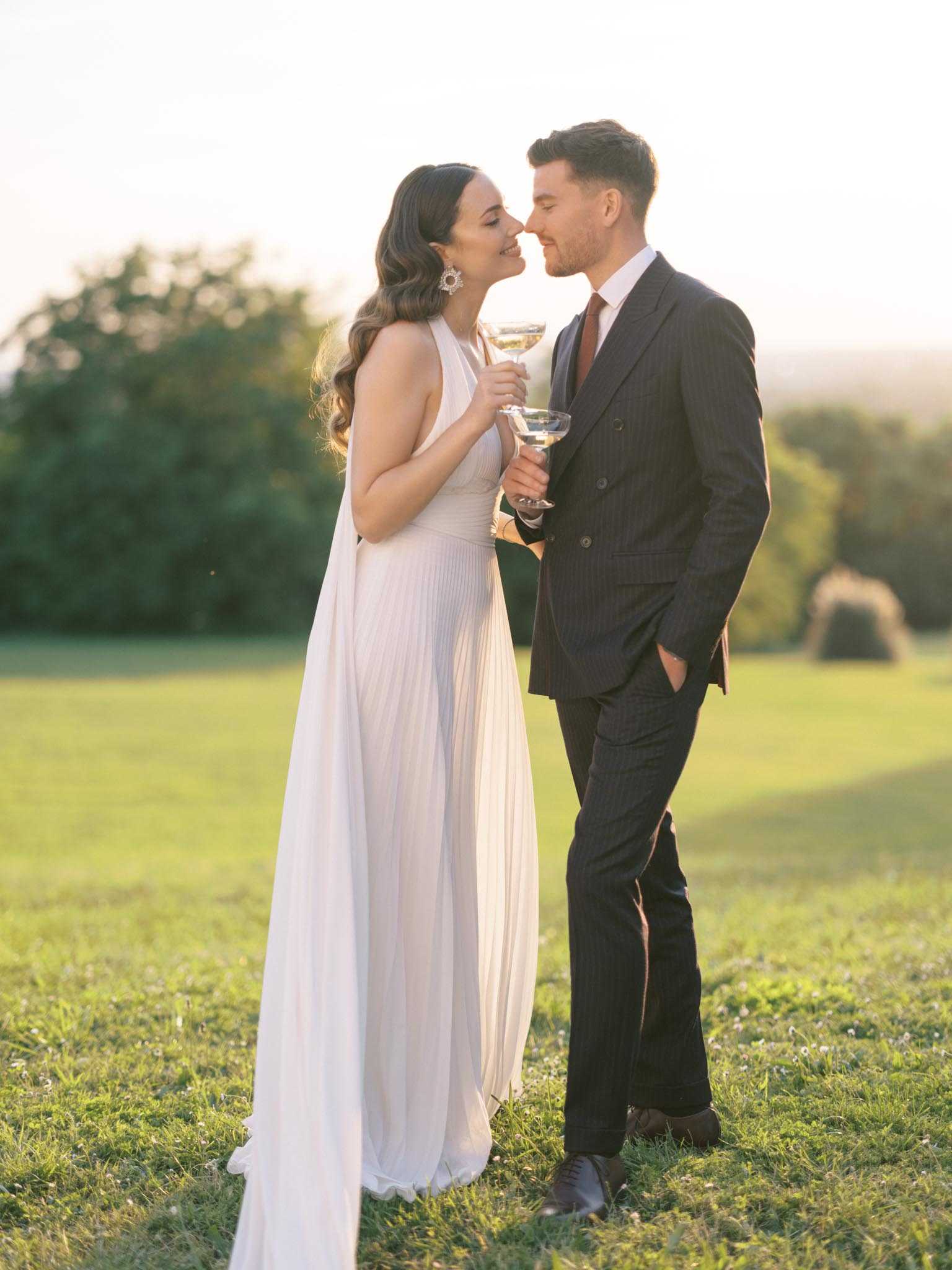 A couple portrait taken outdoors in an open field during golden hour, with warm late-afternoon light backlighting the scene. The bride wears a white halter-neck pleated gown with a flowing cape-style back panel, accessorized with large statement earrings and soft wavy hair worn down; the groom wears a dark navy pinstripe double-breasted suit with a rust-brown tie. The two are leaning in close as if about to kiss while each holding a coupe-style champagne glass. The styling is classic and refined, with a modern edge introduced by the architectural dress silhouette and the groom's pinstripe suit. Full-length couple portrait with a softly blurred open landscape in the background.