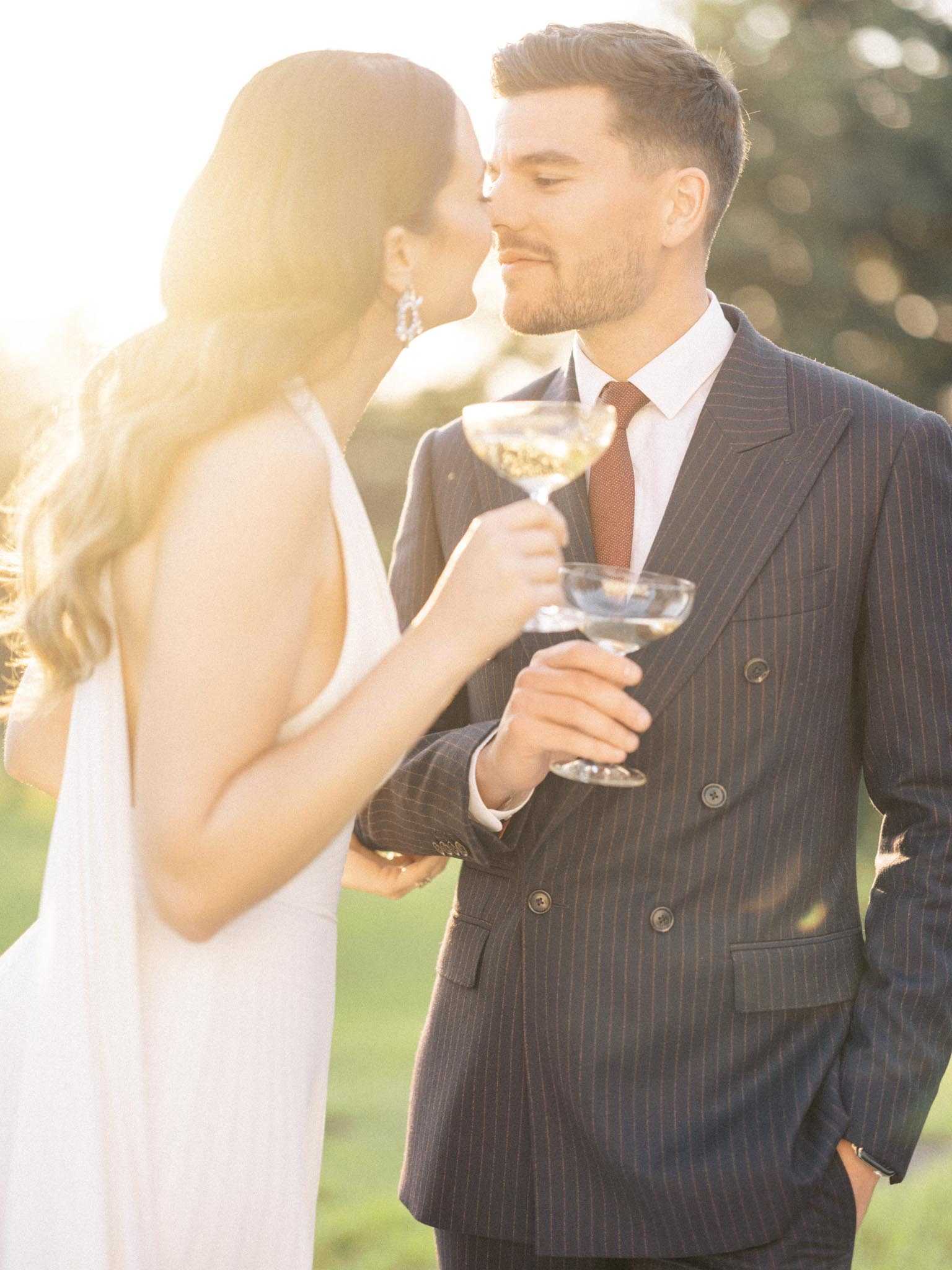 An outdoor couple portrait shot at golden hour, with the bride and groom leaning in to kiss while each holding a coupe champagne glass. The bride wears a sleeveless white dress with an open back and crystal drop earrings, with loose wavy hair. The groom wears a double-breasted navy pinstripe suit with a white shirt and a terracotta-red tie. Strong backlit golden sunlight creates significant lens flare and a warm, hazy glow around the couple. The composition is a medium portrait shot with a shallow depth of field, keeping the background of greenery softly out of focus.