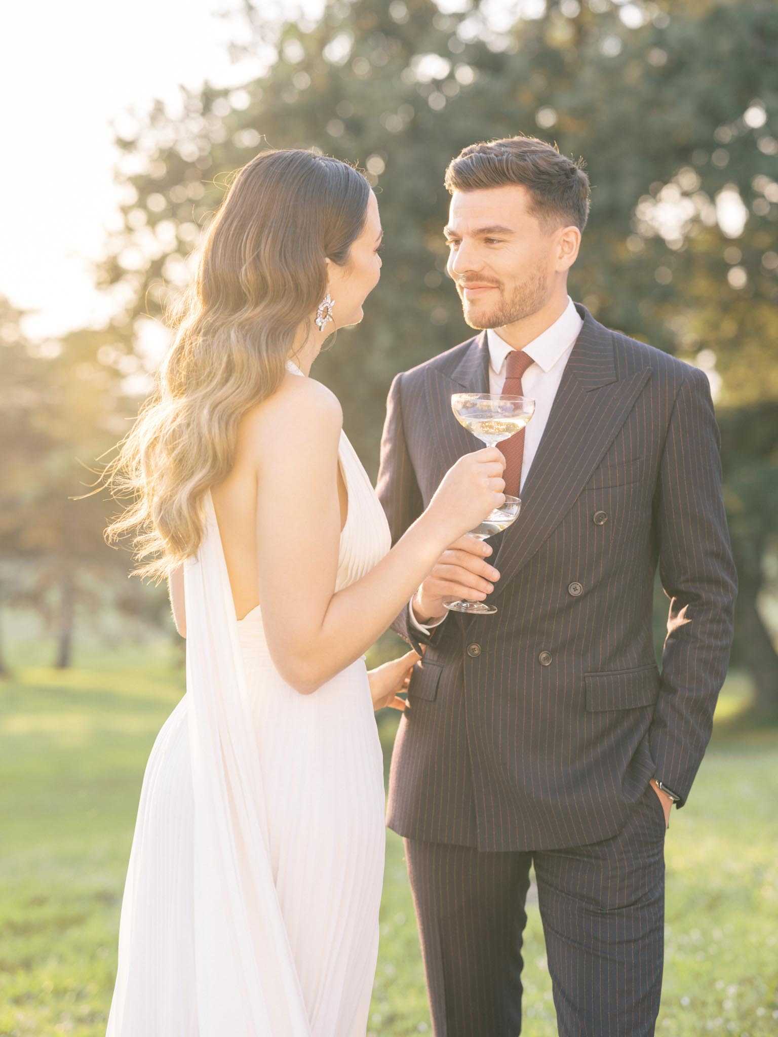 A couple portrait taken outdoors during golden hour, with the bride and groom sharing a champagne coupe toast in a garden or parkland setting. The bride wears a white halter-neck gown with a deep open back and flowing fabric, paired with statement crystal drop earrings, and has long wavy hair. The groom wears a dark navy pinstripe double-breasted suit with a white dress shirt and a burgundy tie. Both are facing each other and smiling, with the groom looking directly at the bride. The composition is a medium portrait shot with a shallow depth of field, creating a soft bokeh background with warm backlit golden tones.