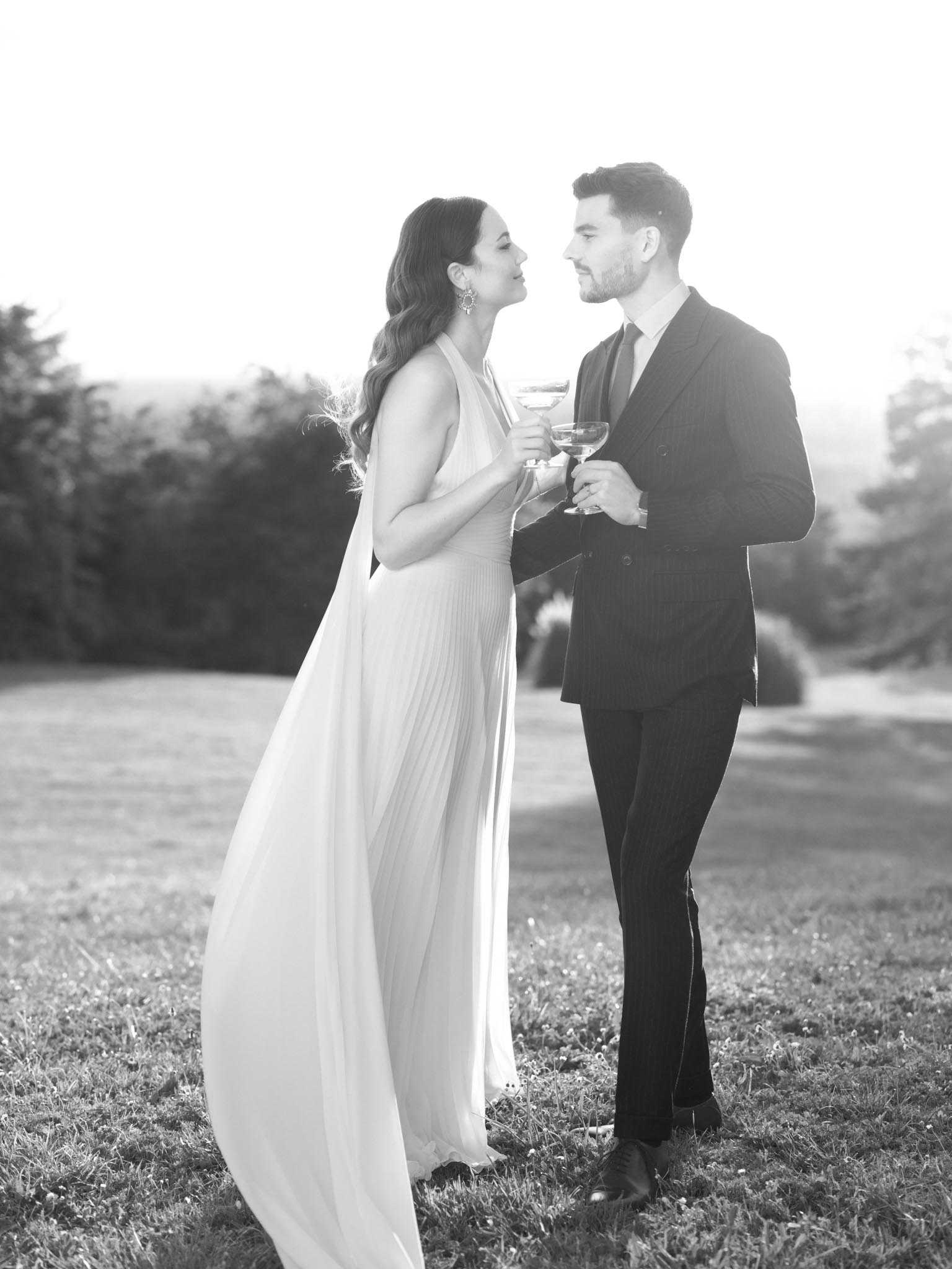 This black-and-white outdoor couple portrait shows a bride and groom standing face-to-face on a lawn, clinking coupe-style champagne glasses and looking at each other closely. The bride wears a halter-neck gown with a flowing, pleated skirt and a cape or train element that drapes behind her, paired with statement drop earrings and loose wavy hair. The groom is dressed in a dark pinstripe double-breasted suit with a tie. The image is lit with strong backlighting that creates bright, blown-out highlights and soft mid-tones, giving the scene a high-contrast, editorial feel. The composition is a full-length portrait shot taken at ground level with the landscape softly out of focus in the background.