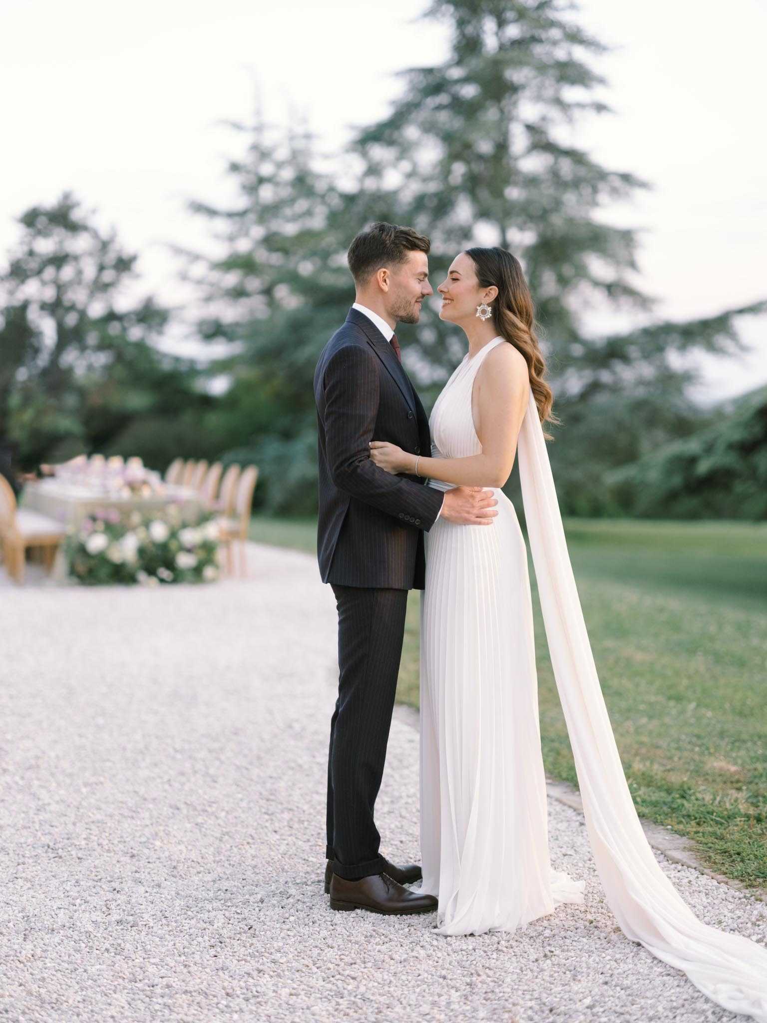 A couple portrait taken outdoors on a gravel path, with the bride and groom facing each other closely and smiling. The groom wears a dark navy pinstripe suit with a burgundy tie, and the bride wears a white pleated sleeveless gown with a long flowing cape train and statement geometric silver earrings, with her hair in loose waves. In the soft-focus background, a long outdoor reception table set with wooden chairs and floral centerpieces in muted tones is visible to the left. The overall styling is modern and clean, with a soft, airy color palette. The shot is a full-length couple portrait with a shallow depth of field.