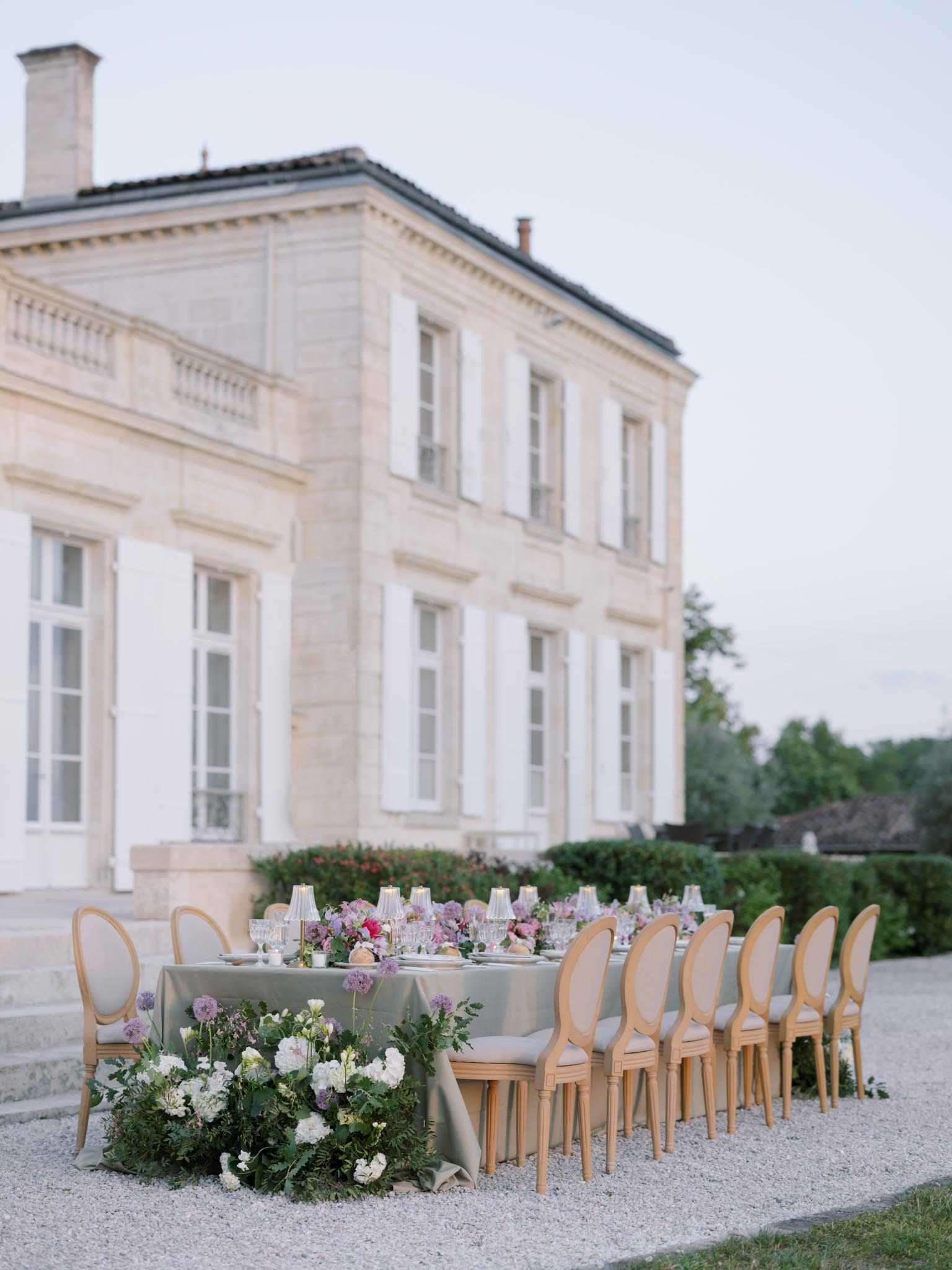 An outdoor wedding reception table is set on a gravel terrace directly in front of a classic French chateau with cream limestone facade and white shuttered windows. The long rectangular table is dressed in a sage green linen tablecloth and lined with natural wood Louis XVI-style oval-back chairs with upholstered seats. A lush floral runner composed of white peonies, cream blooms, allium, and trailing greenery cascades from the near end of the table, while mauve, lilac, and blush florals continue along the center. Small crystal table lamps serve as candlelight accents down the length of the table, accompanied by glassware and place settings. The decor palette combines sage, muted mauve, and natural wood in a classic French style. Wide shot taken at dusk with soft, even light. Potential venue feature image.