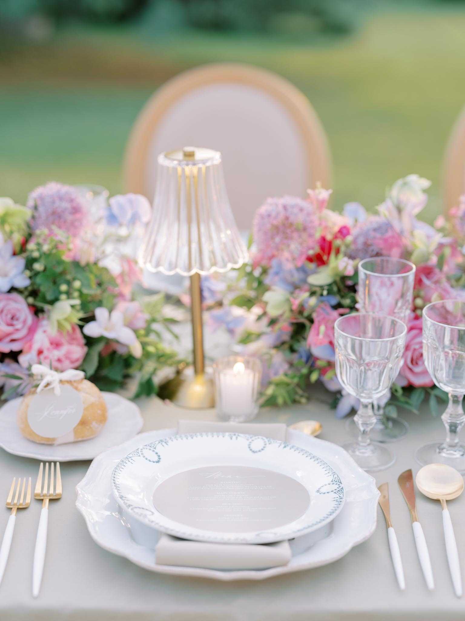 Close-up detail shot of an outdoor wedding reception place setting on a pale grey linen tablecloth. The place setting features a white scallop-edged charger plate with a blue decorative border pattern, topped with a round printed menu card, and a folded grey linen napkin; gold and white-handled flatware is arranged on either side. A small glass and gold table lamp with a ribbed pleated shade serves as an individual place accent, accompanied by a small votive candle. The floral runner consists of pink garden roses, mauve and periwinkle hydrangeas, pale blue sweet peas, and greenery in a pastel, garden-style palette. Crystal pressed-glass goblets in multiple sizes are placed to the right, and a small favor — a wrapped jar tied with white ribbon and a calligraphy tag — sits to the left. A gold Louis-style chair is visible in the background. The overall styling is romantic and French-inspired with a pastel pink, lavender, and blue color palette, gold accents, and vintage-style tableware.
