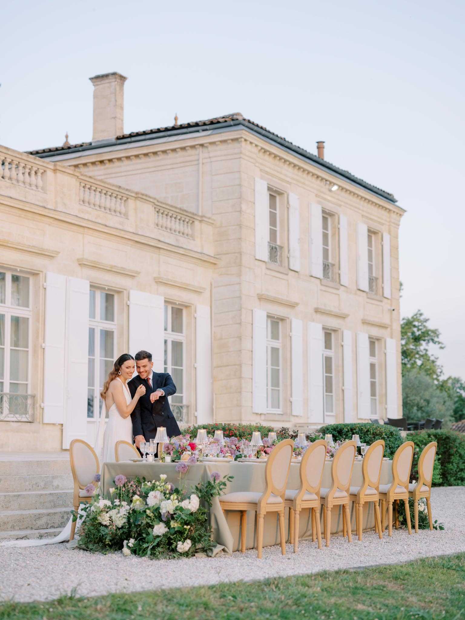 A couple poses beside a long outdoor reception table set on a gravel terrace directly in front of a classic French chateau with limestone facades and white shutters. The bride wears a sleeveless white gown and the groom is dressed in a navy suit; they are leaning close together and smiling downward. The rectangular table is dressed in a sage green linen and lined with gold Louis XVI-style chairs with taupe upholstery, seating approximately 10–12 guests. The tablescape features an abundant low floral runner with ivory dahlias, blush and mauve blooms, greenery and ferns, complemented by tall taper candles in gold holders and glassware. The wide shot captures the full table, the couple, and the full two-storey chateau facade as a backdrop, shot at dusk in soft, warm ambient light. Potential venue feature image.