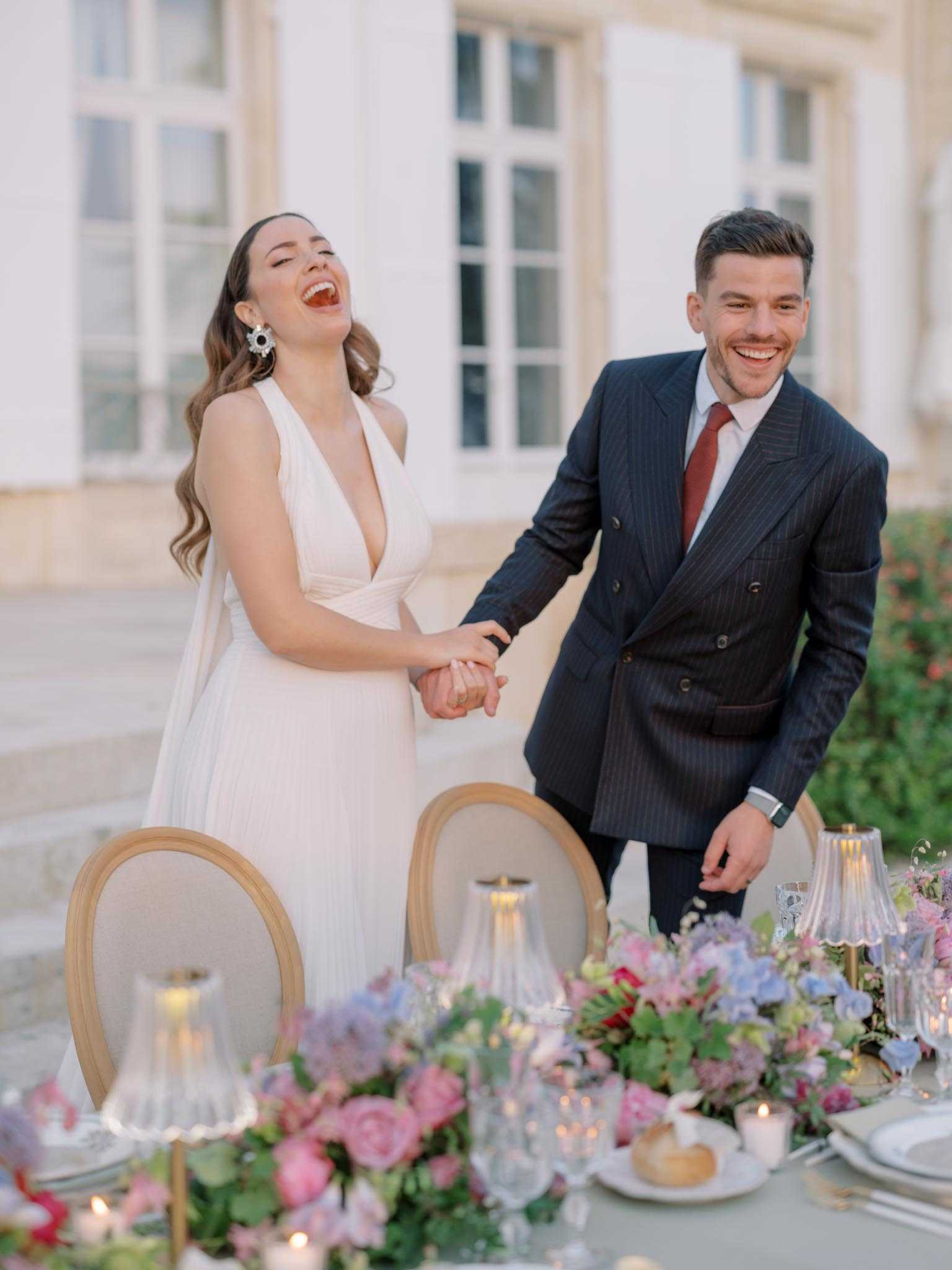 A bride and groom stand together laughing and holding hands beside a decorated outdoor reception table, set against the white facade of a French chateau. The bride wears a deep V-neck ivory pleated gown with statement floral crystal earrings and loose wavy hair, while the groom wears a navy pinstripe double-breasted suit with a burgundy tie. The tablescape in the foreground features lush arrangements of pink garden roses, dusty blue and mauve hydrangeas, and trailing greenery, alongside ribbed glass candle holders with lit candles, crystal glassware, and white plates on a sage green linen tablecloth. The shot is a medium portrait capturing the couple from roughly the waist up, with the table decor artfully blurred in the foreground.
