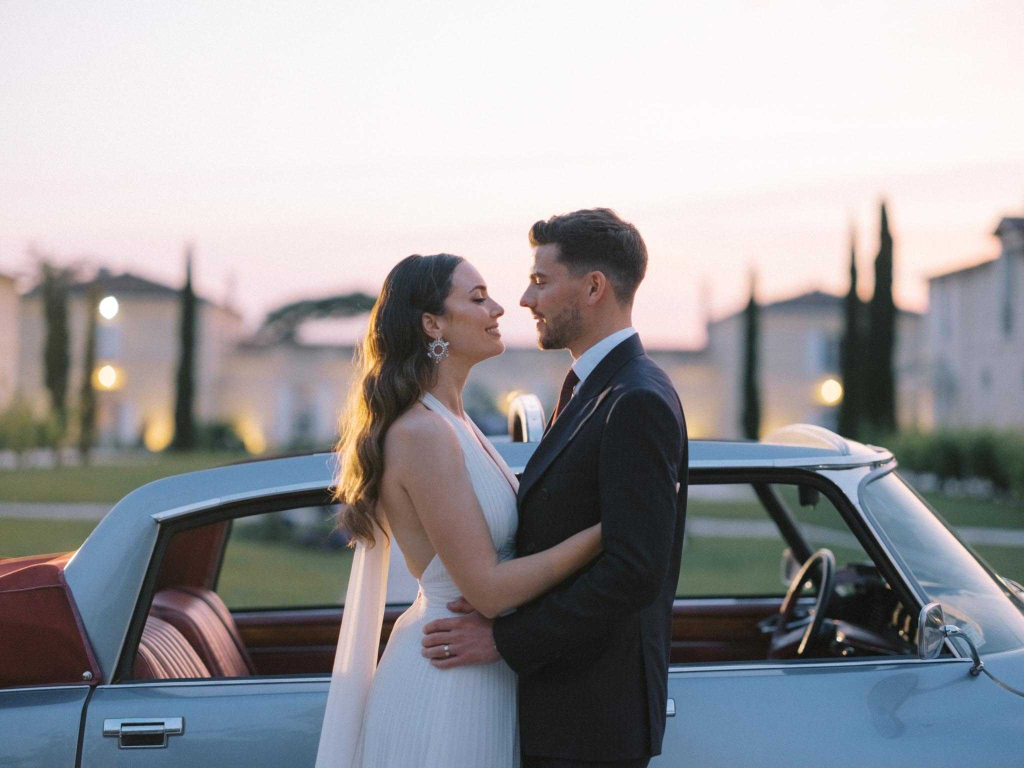 A couple portrait taken outdoors at dusk in front of a vintage pale blue car with red leather interior, likely a Citroën DS, with a château building and tall cypress trees lit by warm ground lights visible in the soft-focus background. The bride wears a white halter-neck dress with a deep V-front and pleated skirt, accessorized with large statement earrings, while the groom wears a dark navy suit with a burgundy tie and white dress shirt. The two stand facing each other in a close embrace, the bride smiling upward toward the groom. The styling has a retro-modern feel, combining the vintage car prop with a clean, minimalist bridal look, and the dusk light gives the image warm golden and soft pink tones. Medium portrait shot, slightly wider to include the car on both sides of the couple.