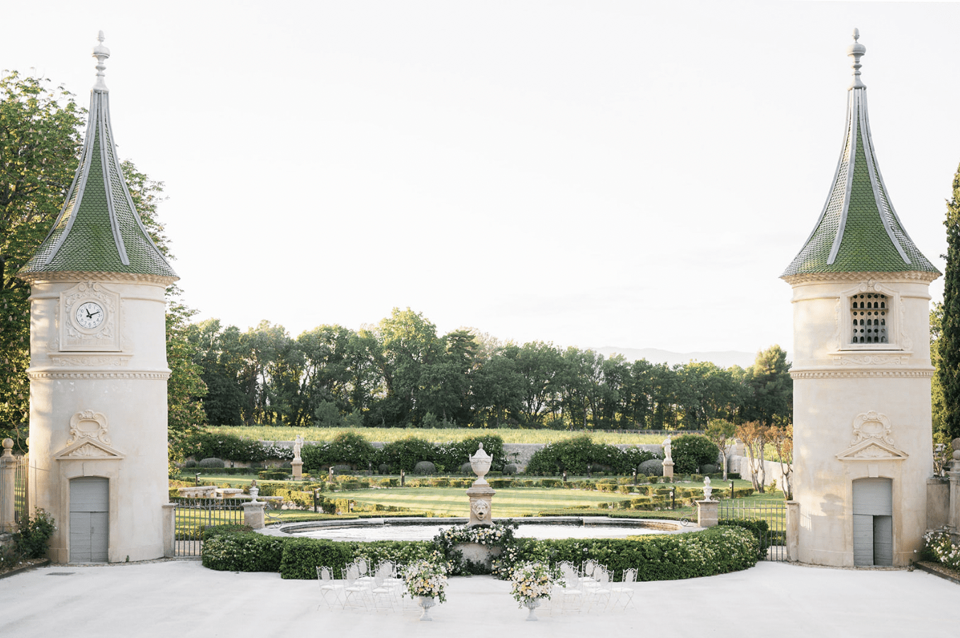 A wide-shot outdoor venue photograph showing a formal French chateau garden set up for a wedding ceremony. Two symmetrical stone pavilions with green tiled conical roofs and decorative carved facades flank the entrance, one featuring a clock face. Between them, a circular stone fountain with a lion-head basin and decorative urn serves as a focal point, surrounded by neatly clipped boxwood hedging. White bistro-style chairs are arranged in a small semicircle around the fountain base, with two large floral arrangements in white urns placed at ground level featuring white, cream, and soft peach blooms. Beyond the fountain, a formal parterre garden extends with manicured hedges, stone statues, and what appears to be additional seating in the distance, backed by a vineyard and tree line. The overall decor palette is white, cream, and soft green with a classic formal French garden aesthetic. Potential venue feature image.