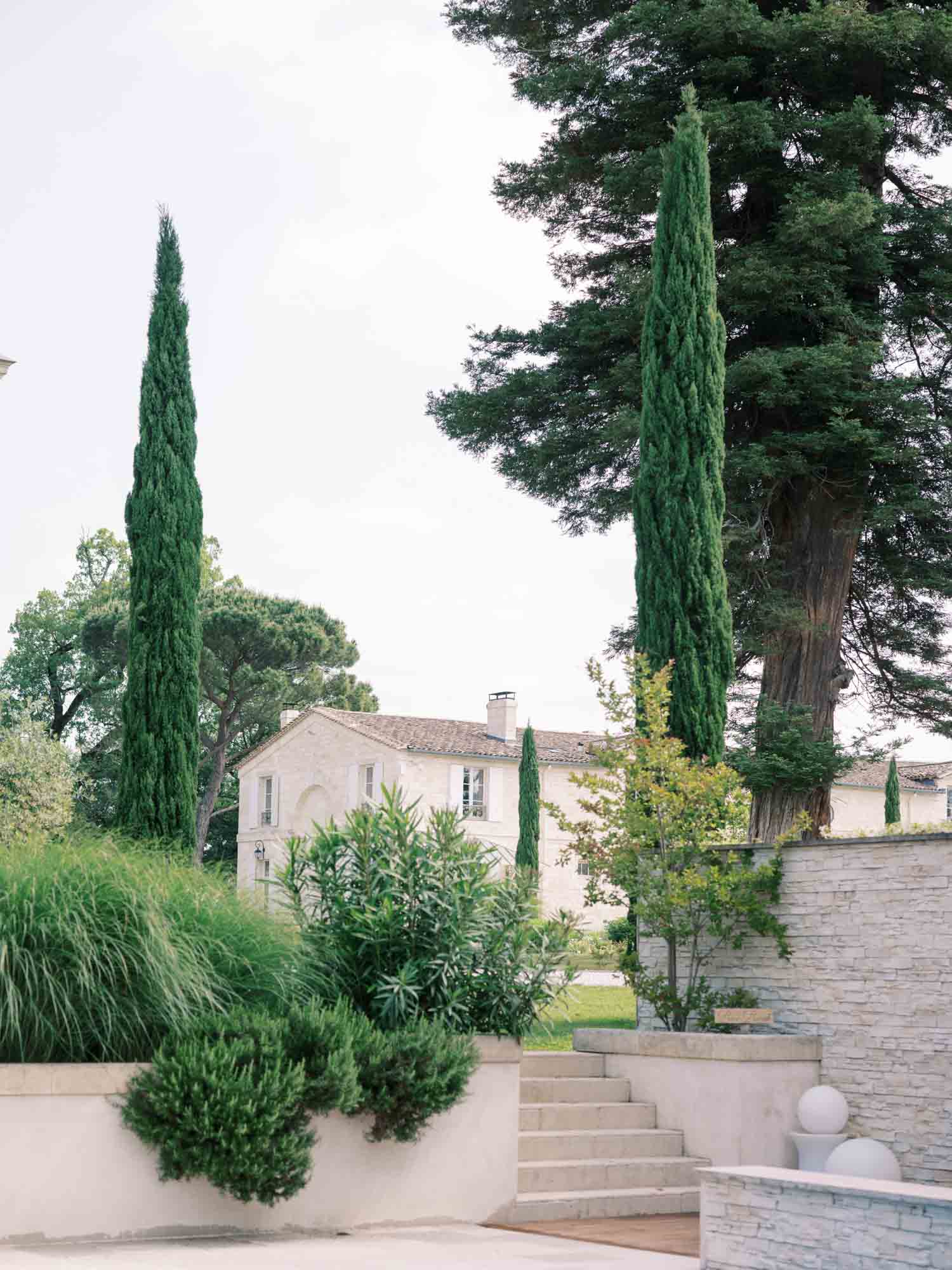 Wide shot of the exterior grounds of a French stone manor house, likely in Provence, featuring a pale limestone facade with arched window details and a terracotta tile roof. The foreground shows a tiered terrace with cream-rendered planter boxes containing clipped shrubs and ornamental grasses, stone steps leading up between levels, and a low dry-stone wall with spherical stone ball finials. Tall Italian cypress trees frame the building on both sides, with a large mature cedar visible to the right. No people are present in the image. Potential venue feature image.