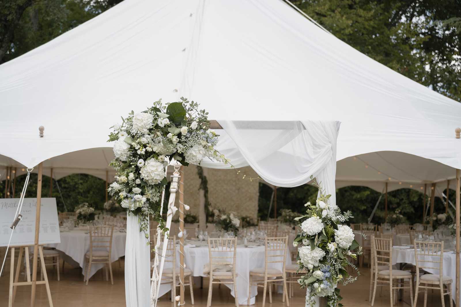 A reception setup shot taken from the entrance of a large white sailcloth tent, framed by a wooden arch decorated with white hydrangeas, white ranunculus, baby's breath, pale blue accents, and trailing eucalyptus foliage. The arch is draped with flowing white fabric. Inside the tent, round tables are covered in white linen and surrounded by natural wood chiavari chairs, with floral centerpieces visible in the background. A seating chart on an easel is positioned to the left of the entrance. The overall decor palette is white, soft blue, and green with a classic, clean aesthetic. Wide shot taken from outside the tent looking inward. Potential venue feature image.