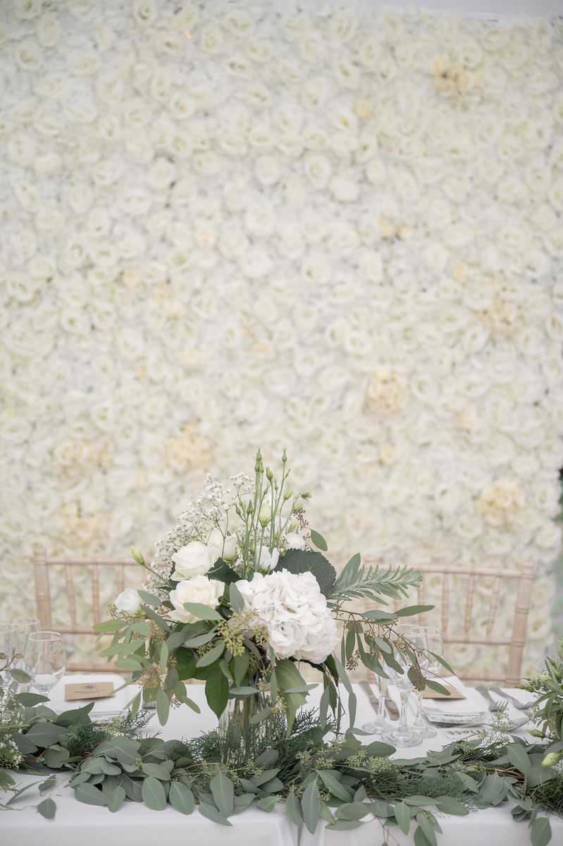 Detail shot of a wedding reception sweetheart or head table setup, styled in an all-white and green palette. The table is dressed with a white linen and features a low centerpiece of white hydrangeas, white roses, lisianthus buds, and baby's breath with trailing eucalyptus and fern greenery; a lush eucalyptus garland runs the length of the table. Place settings include clear glassware, white folded napkins, and small kraft-paper name cards or menus. Two gold chiavari chairs are visible behind the table. The entire arrangement is set against a large floor-to-ceiling flower wall made of densely packed ivory and cream roses, creating a cohesive white-on-white backdrop. The overall styling is classic and clean with a modern garden aesthetic.