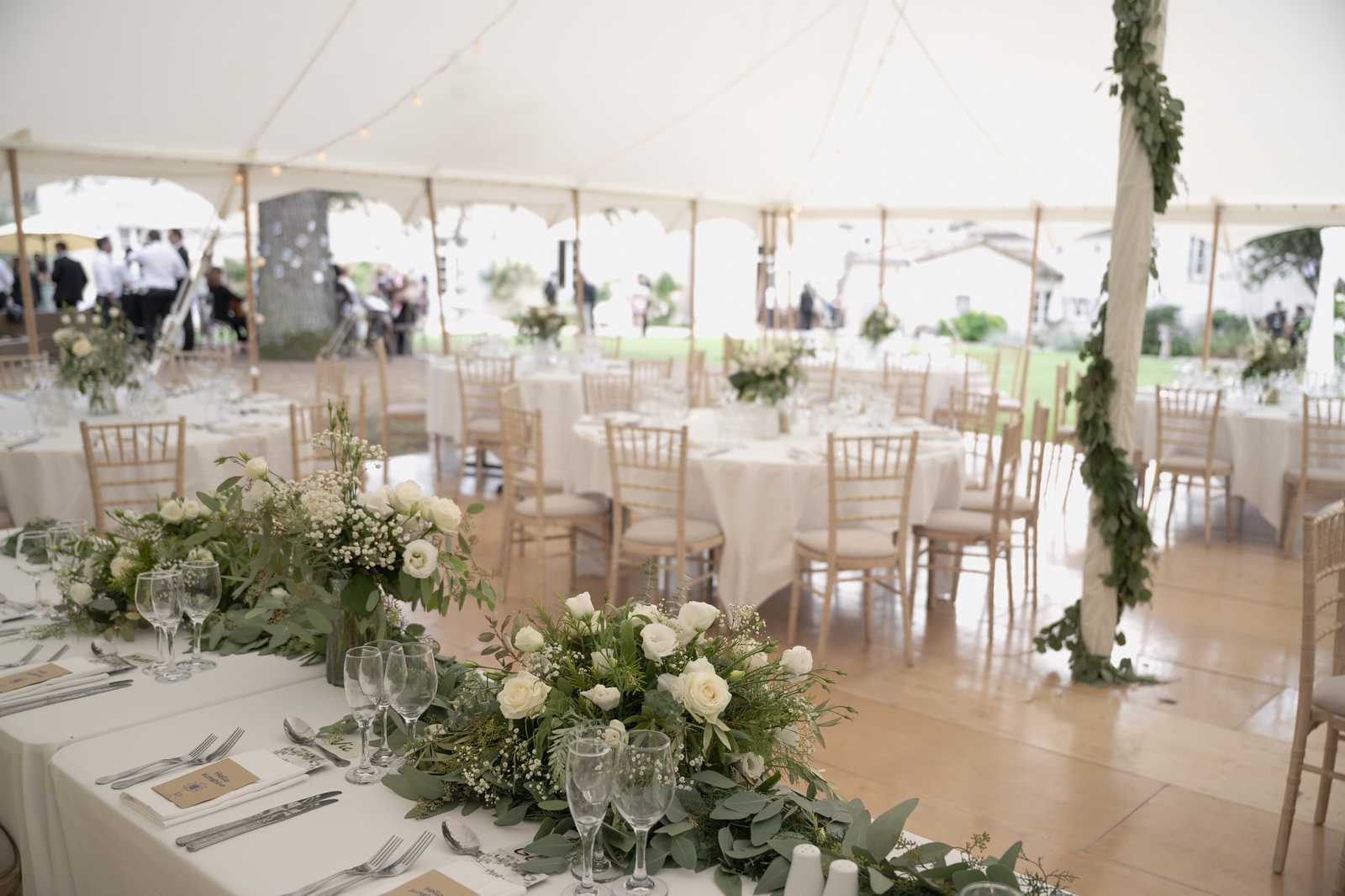 A wedding reception set up inside a large white sailcloth tent with warm-toned wooden flooring and natural wood chiavari chairs. The foreground features a long head table dressed in white linen with a trailing garland of eucalyptus, white roses, baby's breath, and mixed greenery running along its length, alongside place settings with silver cutlery, wine glasses, and kraft paper menu cards. Round guest tables with white linen tablecloths and matching floral centerpieces of white roses and eucalyptus fill the background, while a tent pole in the right foreground is wrapped with lush green ivy. String lights run along the tent ceiling, and guests and staff can be seen mingling in the background near the open sides of the tent. The overall decor palette is white and green with natural wood accents, giving a classic garden-party aesthetic. Wide interior shot.