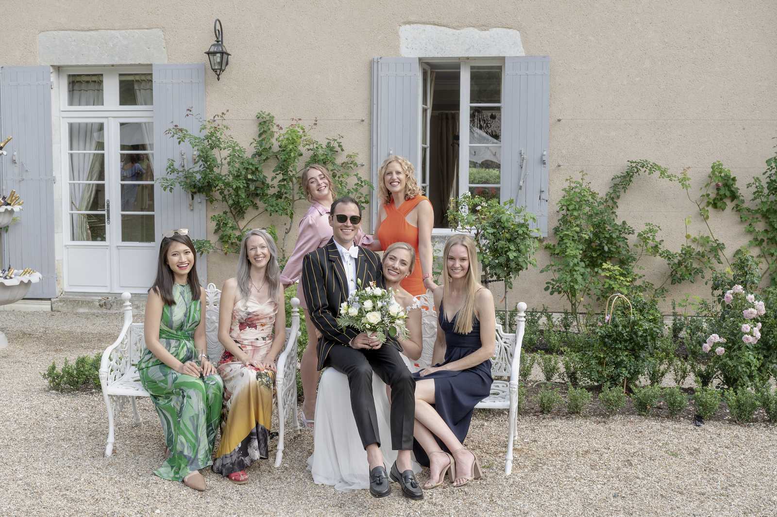 A group portrait taken outdoors in the gravel courtyard of a French country house or small chateau, featuring the couple and five female guests. The groom wears a black blazer with gold pinstripes, black trousers, and loafers with sunglasses, while the bride is in a white wedding dress and holds a bouquet of ivory and pale yellow roses with greenery. The group is arranged across two white ornate cast-iron garden benches, with three women seated and two standing behind. Guest attire includes a green floral maxi dress, a multicolored floral slip dress in ochre and pink tones, a navy slip dress, a blush pink satin top, and an orange sleeveless dress. The building behind features pale blue shutters and climbing roses. Wide group shot taken in natural daylight.