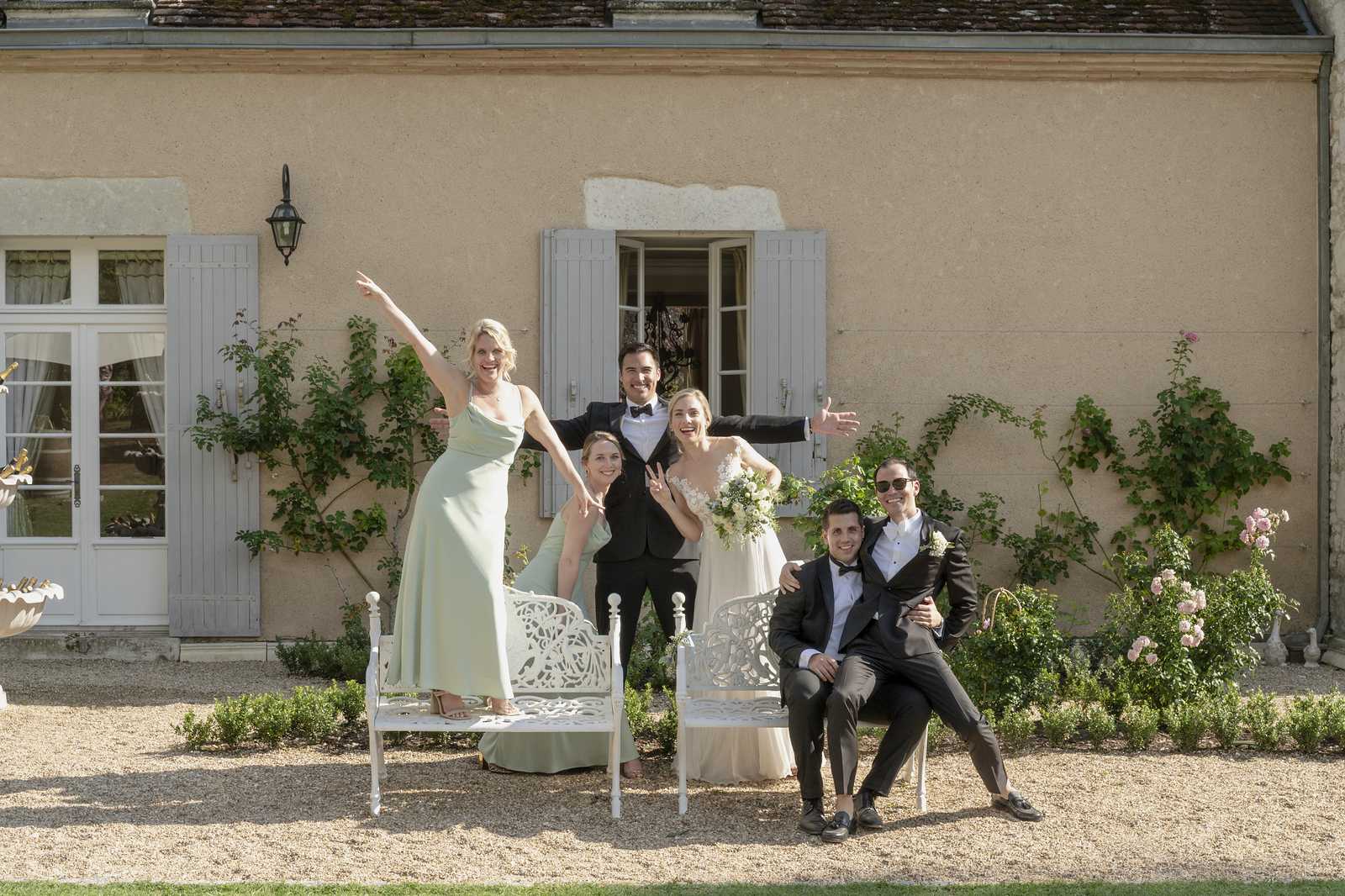 A fun bridal party group portrait taken outdoors on the gravel grounds of a French chateau or manor house, with pale rendered walls, grey shuttered windows, and climbing roses and vines visible on the facade. The group of six includes the couple at the center — the groom in a black tuxedo with bow tie and the bride in a lace-bodice ivory gown holding a white and pale green bouquet — along with two women and two men who appear to be the wedding party, all striking playful, celebratory poses with arms raised and peace signs. The bridesmaids wear sage green floor-length slipdresses, while the groomsmen are dressed in black tuxedos with bow ties; two white ornate cast-iron garden benches are positioned in front of the group. The styling theme is classic French with a soft, neutral palette, and the shot is a wide mid-distance portrait capturing the full group and building facade.