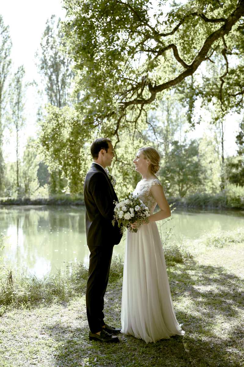 Outdoor couple portrait taken beside a calm pond or small lake, with tall trees and dense foliage in the background. The bride wears a floor-length white chiffon gown with a lace cap-sleeve bodice and her hair styled up, holding a round bouquet of white roses and ranunculus with greenery and small white filler flowers. The groom wears a dark navy or black suit with a white dress shirt. The two are facing each other and smiling, positioned under the branches of a large oak tree. The shot is a full-length portrait taken at a slight distance, capturing both figures in the dappled natural light filtering through the tree canopy.