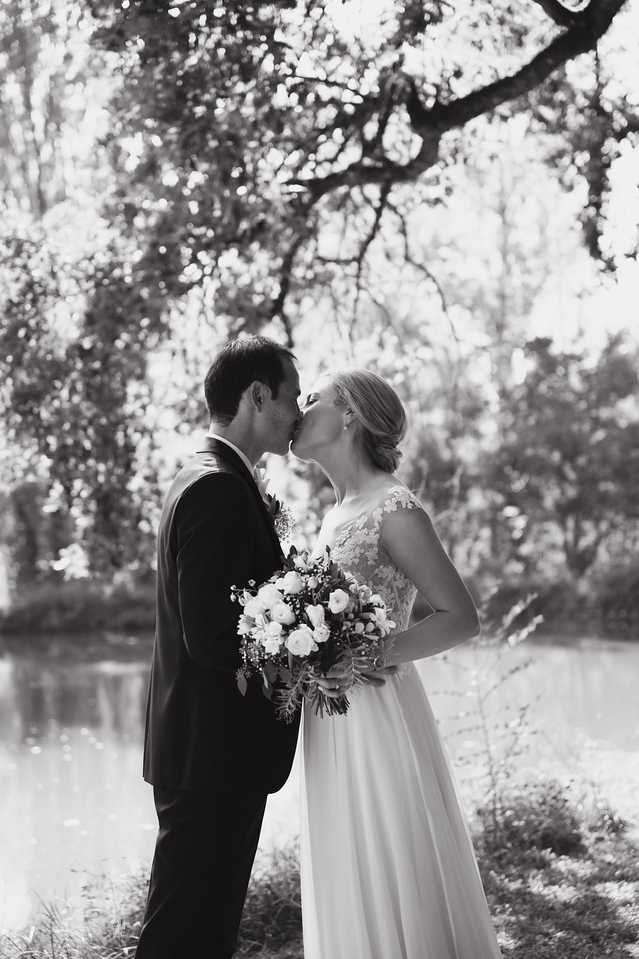 This is a black-and-white couple portrait taken outdoors near a body of water, with trees providing a softly blurred background and bright, diffused natural light creating high contrast between light and shadow. The bride and groom are kissing; she wears a flowing A-line gown with a lace appliqué bodice and cap sleeves, while he wears a dark suit with a boutonniere. The bride holds a full, rounded bouquet featuring what appear to be roses and mixed greenery with darker accent flowers, rendered in mid-tones against the light dress. The composition is a full-length portrait with the couple centered in the frame.