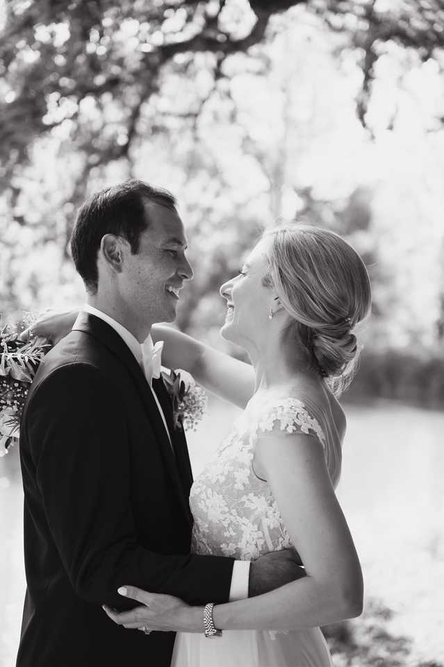 Black-and-white couples portrait taken outdoors, with the bride and groom facing each other, laughing and embracing. The groom wears a dark suit with a bow tie and a floral boutonniere with foliage, while the bride wears a lace cap-sleeve wedding gown with floral appliqué detailing and has her hair pulled back in a low chignon with drop earrings and a bracelet. The bride's hand rests gently on the groom's chest as he holds her waist. High-contrast bright tones in the background create a soft, light-filled backdrop with blurred tree canopy. The composition is a mid-length portrait shot.