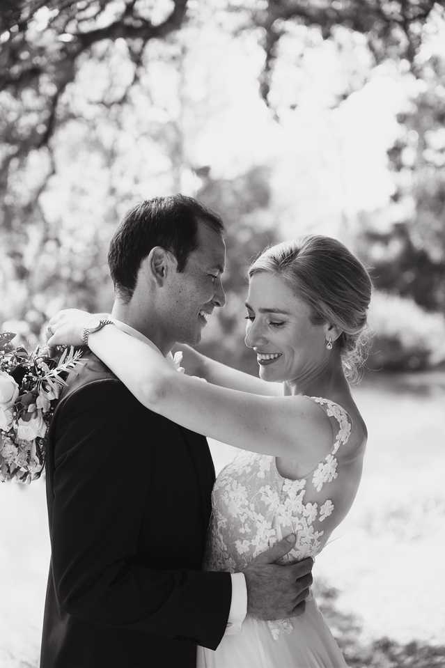 This is a black-and-white couple portrait taken outdoors, with a softly blurred tree canopy forming a bright, high-key background. The bride and groom face each other closely, both smiling and laughing; the bride has her arm draped around the groom's shoulder while he holds her at the waist. The bride wears a lace-appliqué gown with a scoop neckline and carries a bouquet visible at the left edge of the frame, featuring what appear to be full blooms and foliage. Her hair is styled in a low updo and she wears drop earrings. The groom is in a dark suit. The image is rendered in soft contrast with bright highlights in the background, giving it an airy, light tone. Close-up portrait composition, waist-to-head framing.