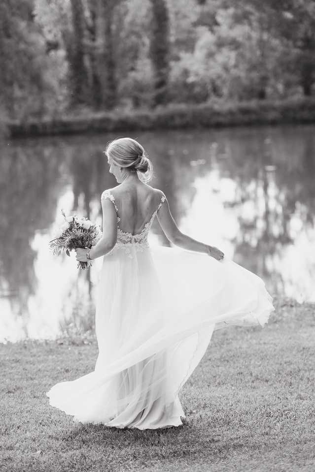 This is a black-and-white bridal portrait shot outdoors beside a reflective pond or lake. The bride is photographed from behind, mid-turn, with the skirt of her flowing chiffon gown caught in motion and billowing outward. Her dress features a low open back with lace appliqué detailing along the bodice and cap-sleeve straps. She wears her hair in a low updo and holds a medium-sized bouquet with varied textures visible in the mid-tones. The image is a full-length portrait with soft contrast, the water and tree reflections in the background rendered in gentle grey tones. The composition emphasizes the movement of the dress and the open back detailing.