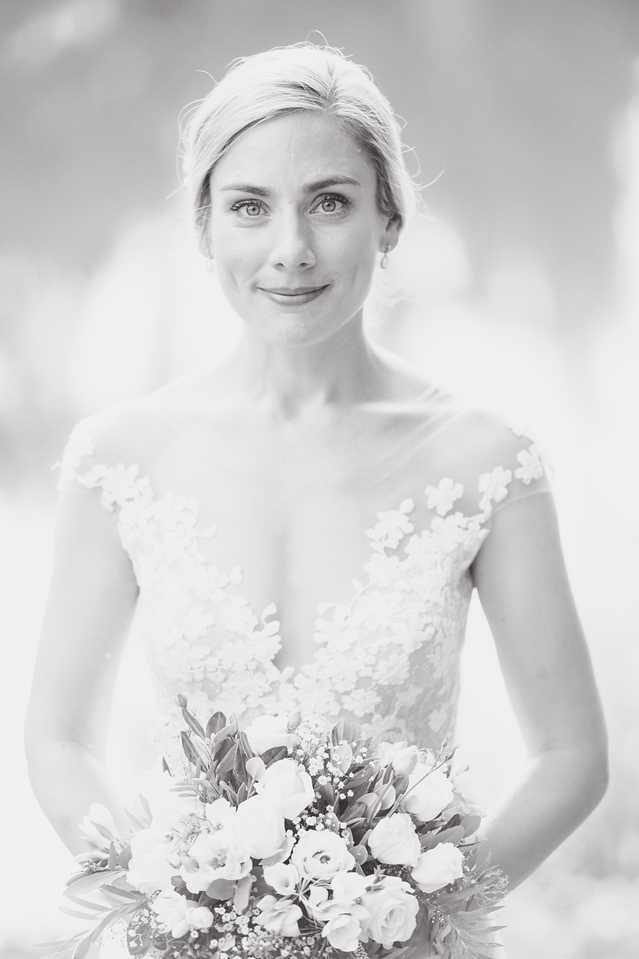 This is a black-and-white bridal portrait shot in a close-up style with a softly blurred background and bright, airy tones with gentle contrast. The bride faces the camera directly with a calm expression, her hair pulled back in an updo with soft pieces framing her face, and she wears small stud earrings. Her gown features a deep V-neckline with a sheer illusion bodice covered in three-dimensional floral appliqués and cap sleeves. She holds a bouquet composed of roses, ranunculus, small lily buds, baby's breath, and trailing foliage, visible in the lower portion of the frame.