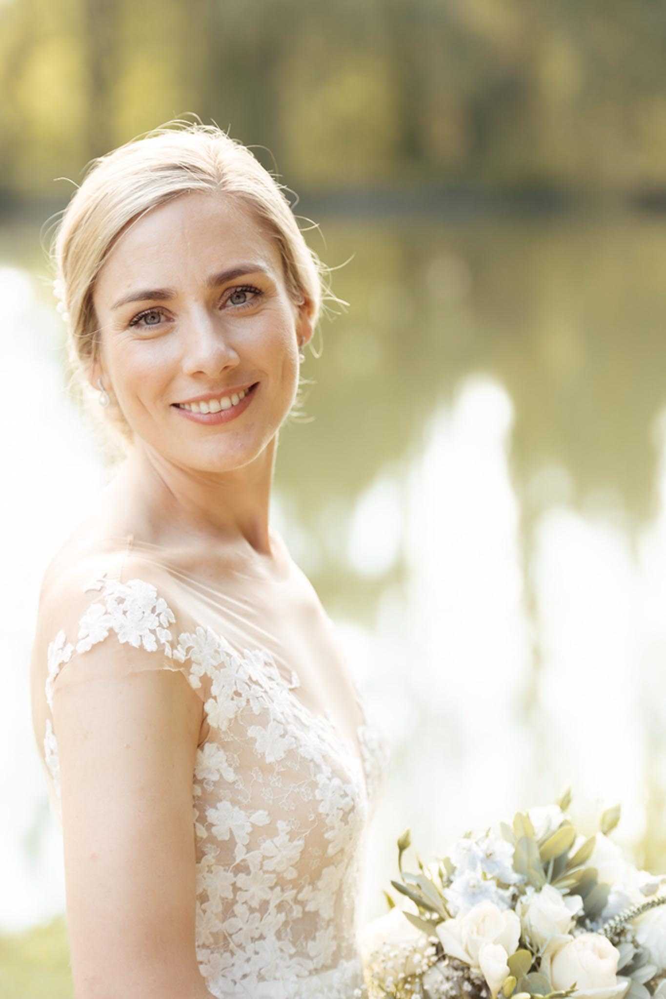 A close-up portrait of a bride outdoors, photographed in front of a softly blurred body of water. She wears a cap-sleeve lace wedding dress with floral appliqué over a nude/blush underlay, and her blonde hair is pulled back in an updo with soft face-framing pieces. She holds a bouquet featuring ivory peonies or ranunculus, pale blue flowers, eucalyptus, and dusty miller. The styling is classic and delicate, with a natural, warm light giving the image a soft, bright tone.