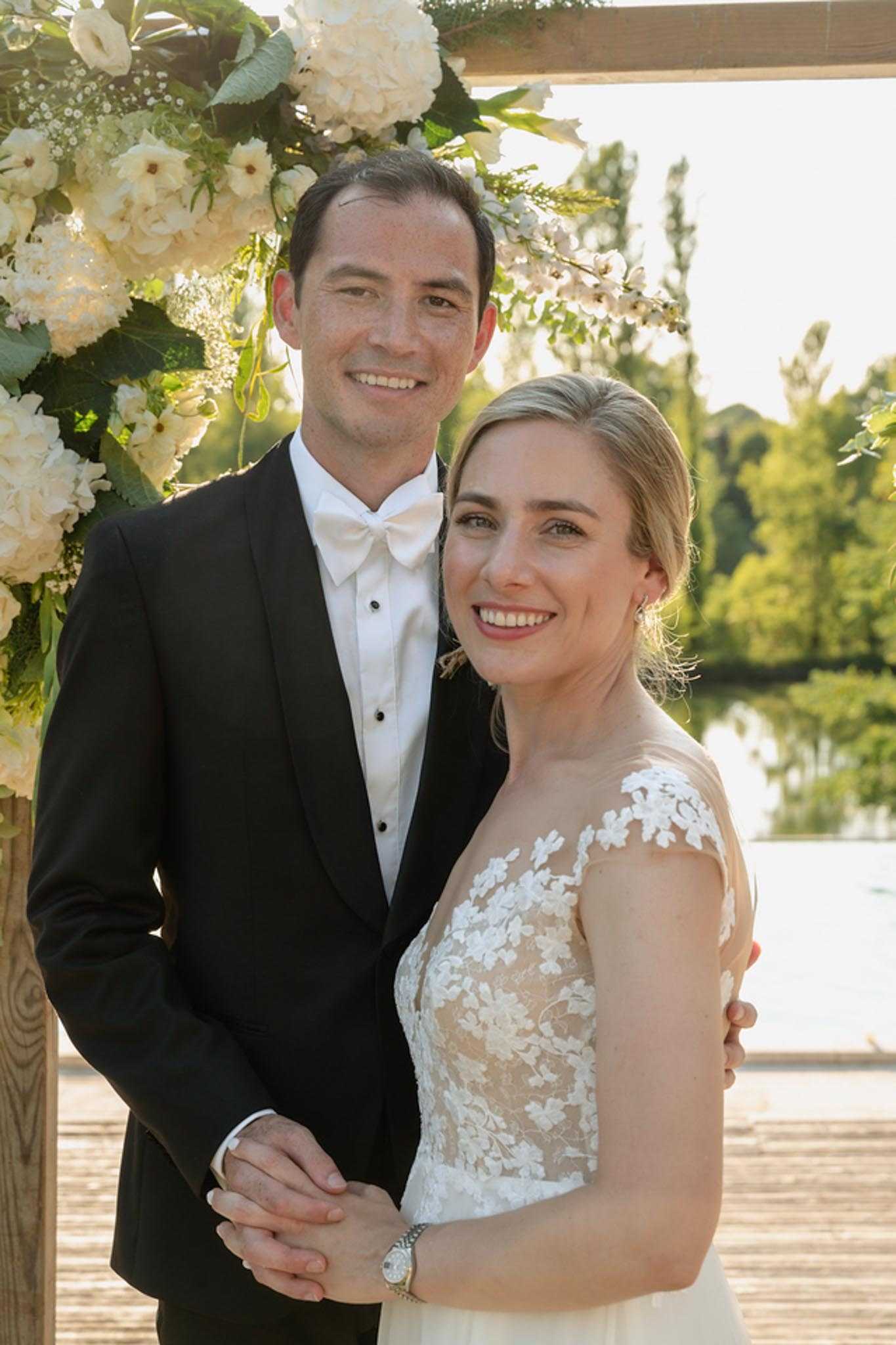 A couple portrait taken outdoors on a wooden deck or dock beside a lake or pond, framed by a wooden arch decorated with cream hydrangeas, white roses, baby's breath, and green eucalyptus foliage. The groom wears a black tuxedo with a white dress shirt, black studded buttons, and a white bow tie, while the bride wears an ivory lace gown with a sheer illusion neckline and floral lace appliqué over the bodice and cap sleeves, her blonde hair pulled back. The overall decor palette is white and ivory with greenery, consistent with a classic, formal styling approach. The image is a medium close-up portrait shot in warm golden-hour light.