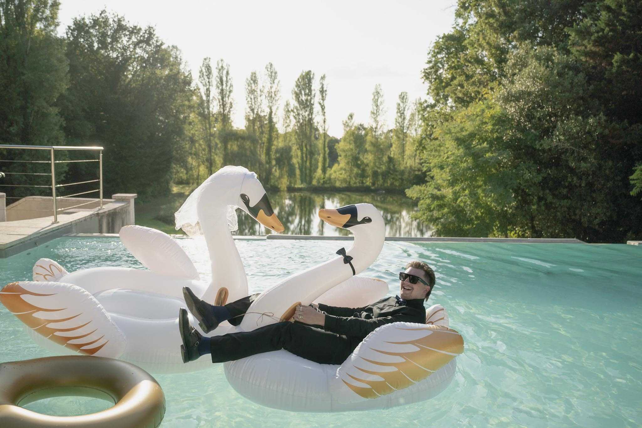 A groom dressed in a black suit with a black bow tie and sunglasses reclines laughing on a large white and gold inflatable swan pool float, with a second white swan float beside him and a gold ring float visible in the foreground. The setting is an outdoor infinity pool overlooking a calm reflective pond surrounded by tall trees, giving the scene a relaxed, playful feel. This appears to be a candid or lighthearted portrait moment, likely during the cocktail hour or reception. The composition is a medium-wide shot taken at pool level, capturing both the groom's full figure and the expansive poolside setting.