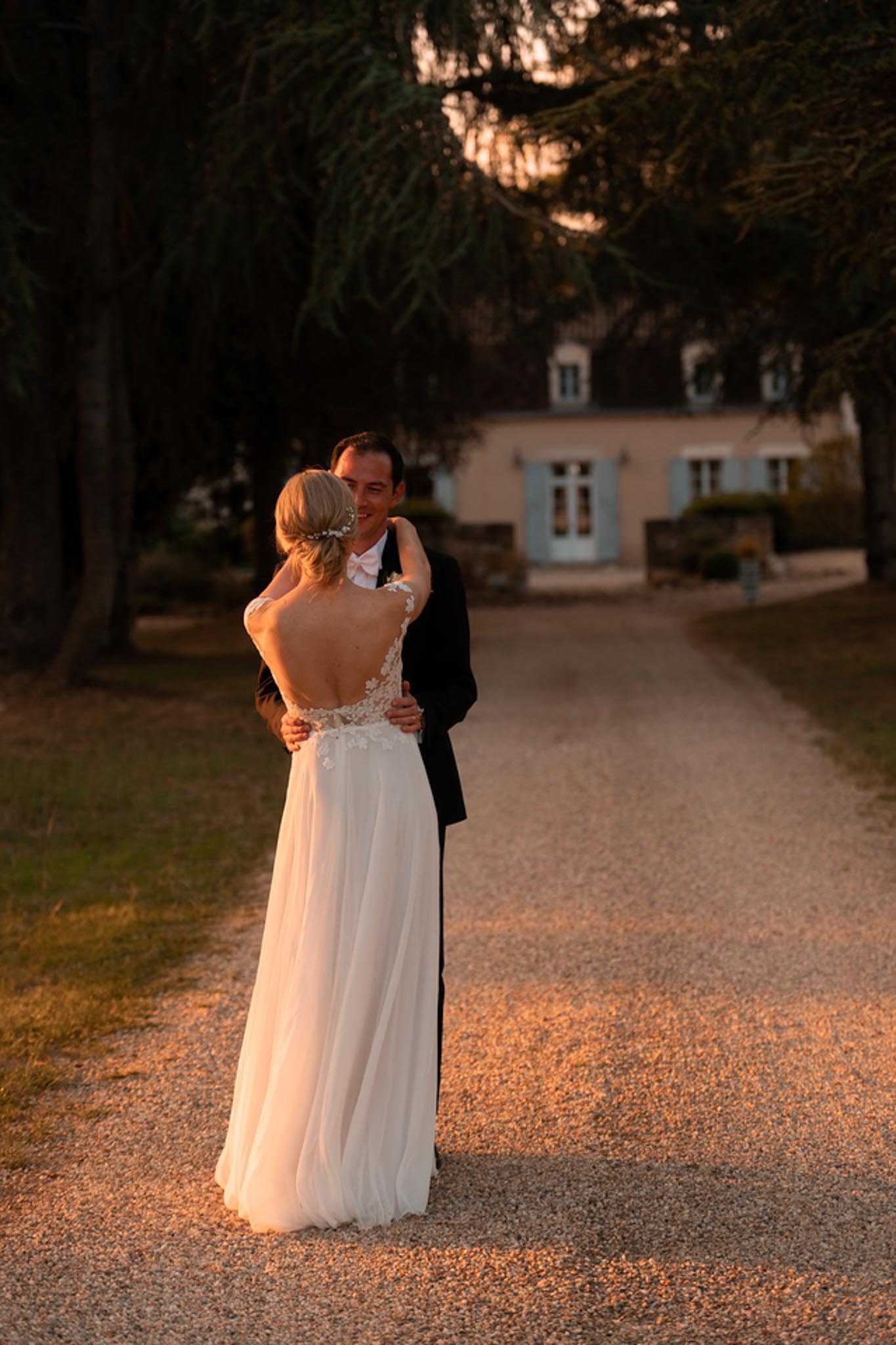 A couple portrait taken outdoors on a gravel driveway leading to a French country manor house with pale pink rendering and pale blue shutters, photographed during golden hour with warm amber light bathing the scene. The bride wears a backless ivory tulle gown with floral lace appliqué across the open back and a small floral hair accessory, her blonde hair styled in an updo; the groom wears a dark navy suit with a blush pink bow tie. The two are embracing and facing each other in a mid-shot framed from behind, with the manor visible and softly out of focus in the background. The overall styling is classic and romantic, shot in a medium portrait orientation. Potential venue feature image.
