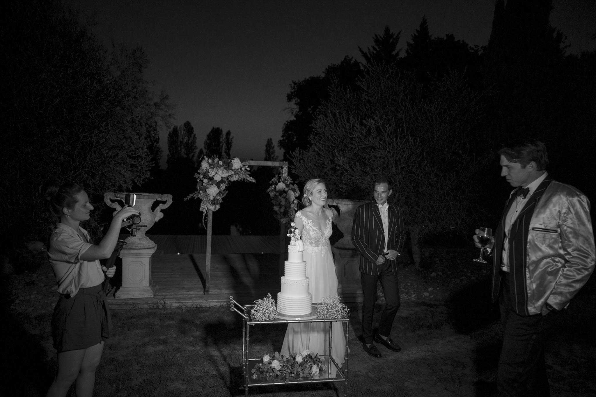 A black-and-white image of an outdoor cake-cutting moment taking place at night in what appears to be a garden or chateau grounds. The bride, wearing a lace V-neck gown with a flowing skirt, stands beside a four-tier white wedding cake topped with a figurine topper, displayed on a gold metal cart surrounded by small floral arrangements at its base. A floral arch with loose blooms is visible in the background alongside large stone urns. Two male guests stand to the right — one in a pinstripe blazer and one in a satin-finish jacket with a bow tie holding a wine glass — while a videographer on the left films the moment using a smartphone on a stabilizer rig. The image has high contrast with deep shadows due to low ambient light, giving it a dark, moody tonality. Wide shot capturing all four people and the full cake display.