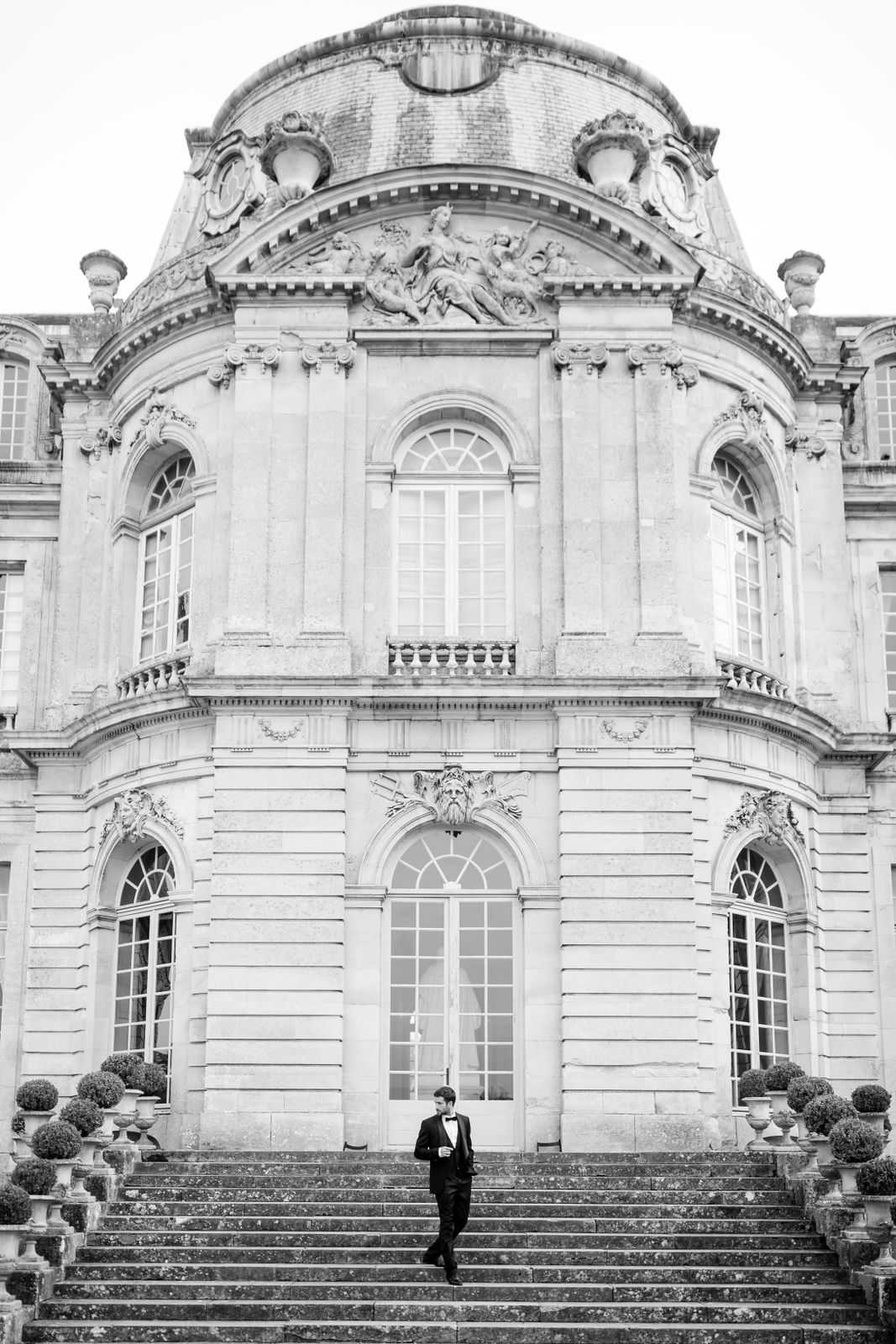 This black-and-white portrait shows a groom dressed in a dark tuxedo with a bow tie descending the grand exterior staircase of a French chateau. The building features classical French Baroque architecture with a domed central pavilion, ornate stone carvings, sculptural relief panels, arched windows, and a decorative balustrade, flanked by rows of clipped topiary shrubs in stone urns. The groom is centered in the lower third of the frame, with the full chateau facade filling the background in high contrast tones. The composition is a wide vertical shot that emphasizes the scale of the architecture relative to the single figure. Potential venue feature image.