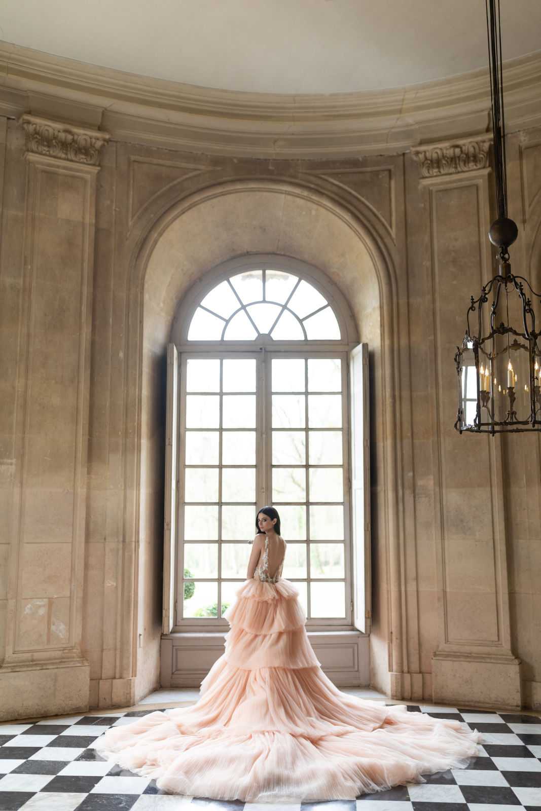 A bridal portrait taken indoors in what appears to be a French chateau or palace interior, featuring a bride standing with her back partially turned toward the camera in front of a tall arched window with fanlight detailing. The bride wears a blush peach tiered tulle ball gown with a deeply open back featuring floral appliqué detailing and an extended cathedral-length train that fans across the black-and-white checkered marble floor. A wrought-iron candle chandelier hangs to the right of the frame. The room has ornate carved stone paneling and curved walls, consistent with 17th or 18th-century French classical architecture. The composition is a wide portrait shot, backlit by soft natural light from the window, emphasizing the volume and sweep of the gown's train. Potential venue feature image.