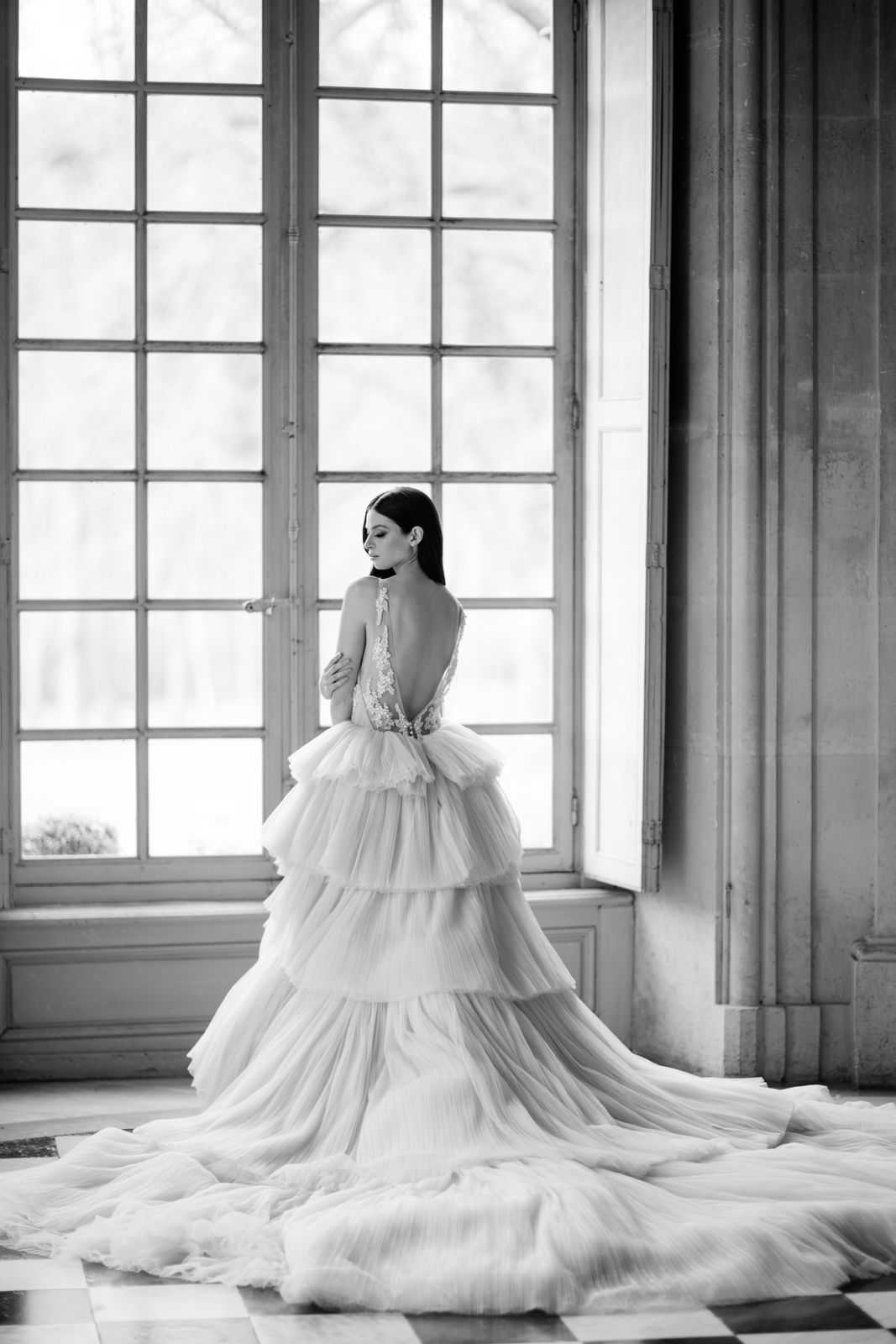 This is a black-and-white bridal portrait shot indoors in what appears to be a grand chateau or palace interior, with tall multi-pane French windows flooding the room with natural light. The bride stands with her back to the camera, looking over her shoulder, wearing a dramatic tiered tulle ballgown with a deep open back, lace appliqué detailing along the spine, and an extended train that spreads across the checkered marble floor. Her dark hair is worn straight and sleek, and she wears a choker-style necklace at her throat. The image is high-contrast black and white, emphasizing the volume and layered texture of the gown's multiple tulle tiers against the bright window light and the ornate stone architectural detailing of the room. The composition is a three-quarter full-length portrait with the gown's train as a central visual element.