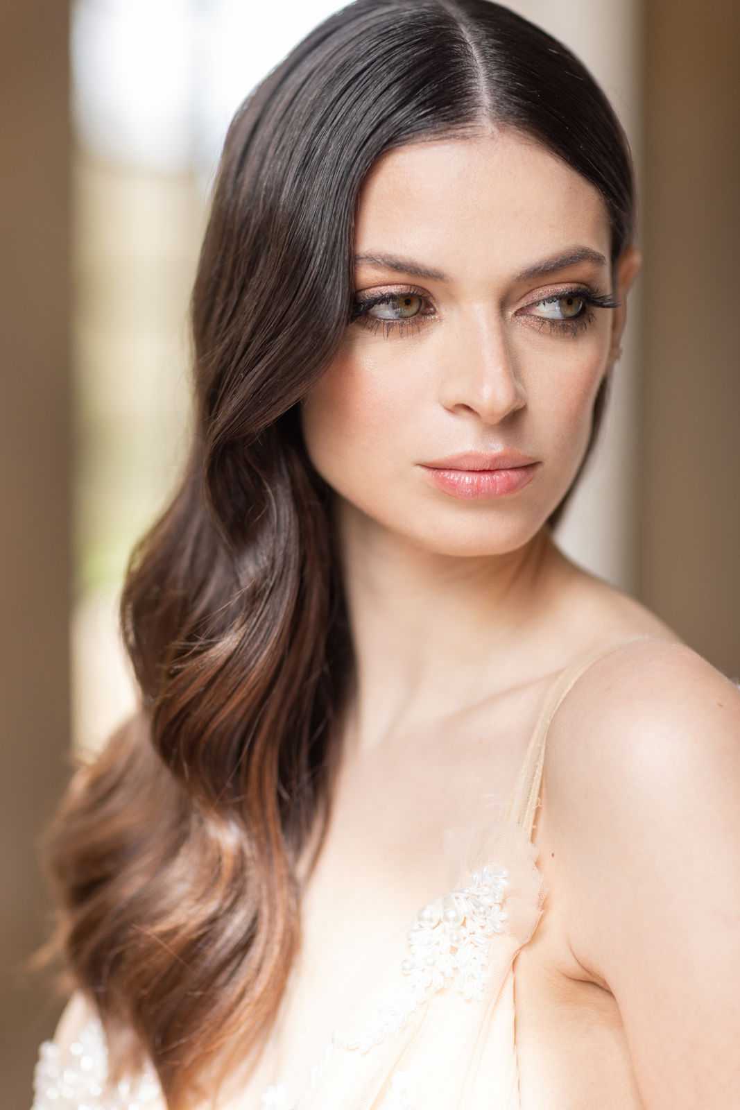 Close-up portrait of a bride during the getting-ready phase, shot indoors with a softly blurred background suggesting large windows or doorways. The bride has long dark brown hair styled in loose side-swept waves and wears bridal makeup including defined brows, smoky eye shadow, extended lashes, and a nude-pink lip. She is wearing what appears to be a blush or champagne slip-style wedding gown or robe with thin spaghetti straps and delicate beaded lace detailing at the chest. The composition is a tightly framed head-and-shoulders portrait with shallow depth of field, emphasizing the hair and makeup styling.