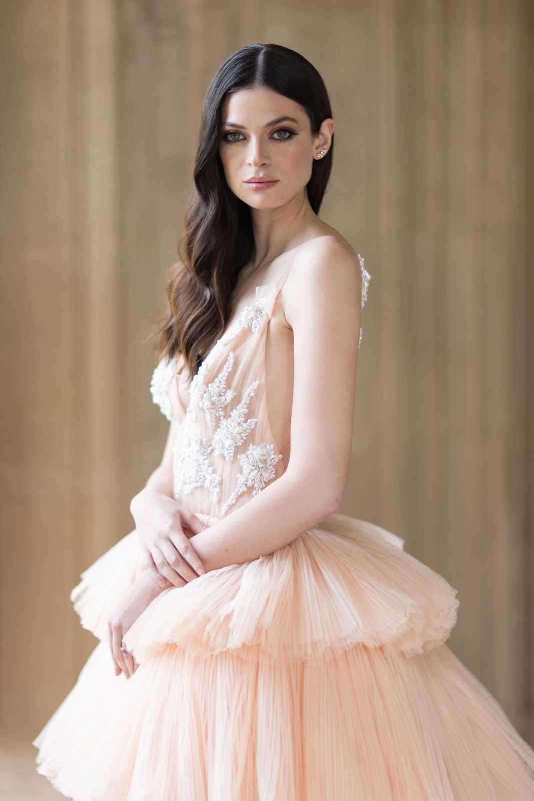 A close-up portrait of a bride wearing a strapless blush peach tulle ball gown with white floral lace appliqué detailing on the bodice and tiered, pleated skirt layers. The bride has dark brown wavy hair swept to one side, defined smoky eye makeup, and wears small crystal or diamond stud earrings. She is posed with her hands clasped in front, facing slightly to the side while looking directly at the camera. The background is softly blurred with warm golden tones, suggesting an indoor setting with architectural paneling or drapery. The overall styling is formal and fashion-forward, with the non-traditional colored gown as the focal point.