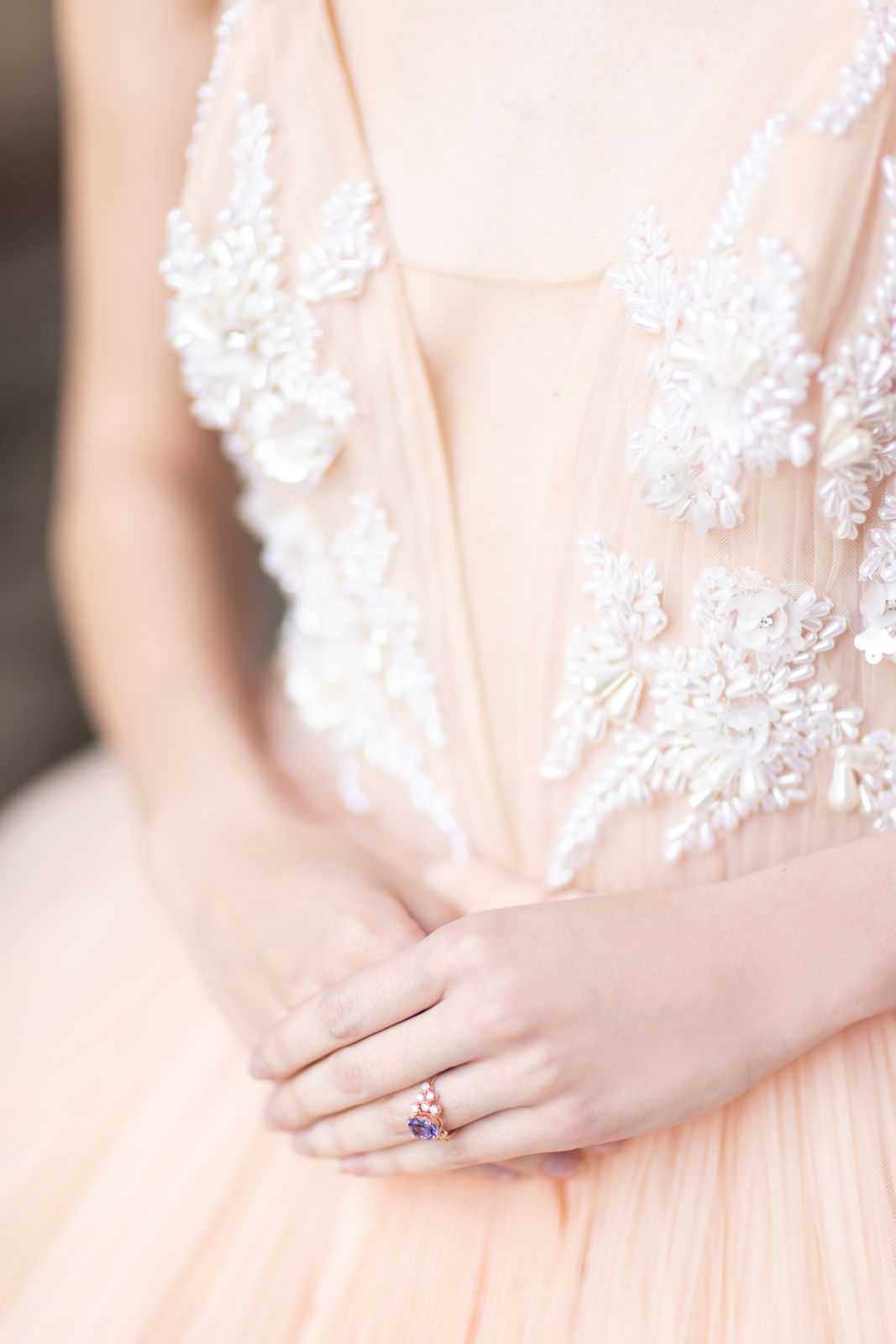 A close-up detail shot of a bride's torso and folded hands, highlighting two key styling elements: a blush pink bridal gown with a sheer bodice heavily embellished with white and ivory beaded floral appliqués and pearl-like beading, and a rose gold engagement ring featuring a blue-purple oval gemstone (likely tanzanite or sapphire) accented by small white stones. The skirt of the gown is pleated blush tulle. The composition is a tight detail portrait with intentional shallow depth of field, keeping the dress embellishment and ring as the focal points.