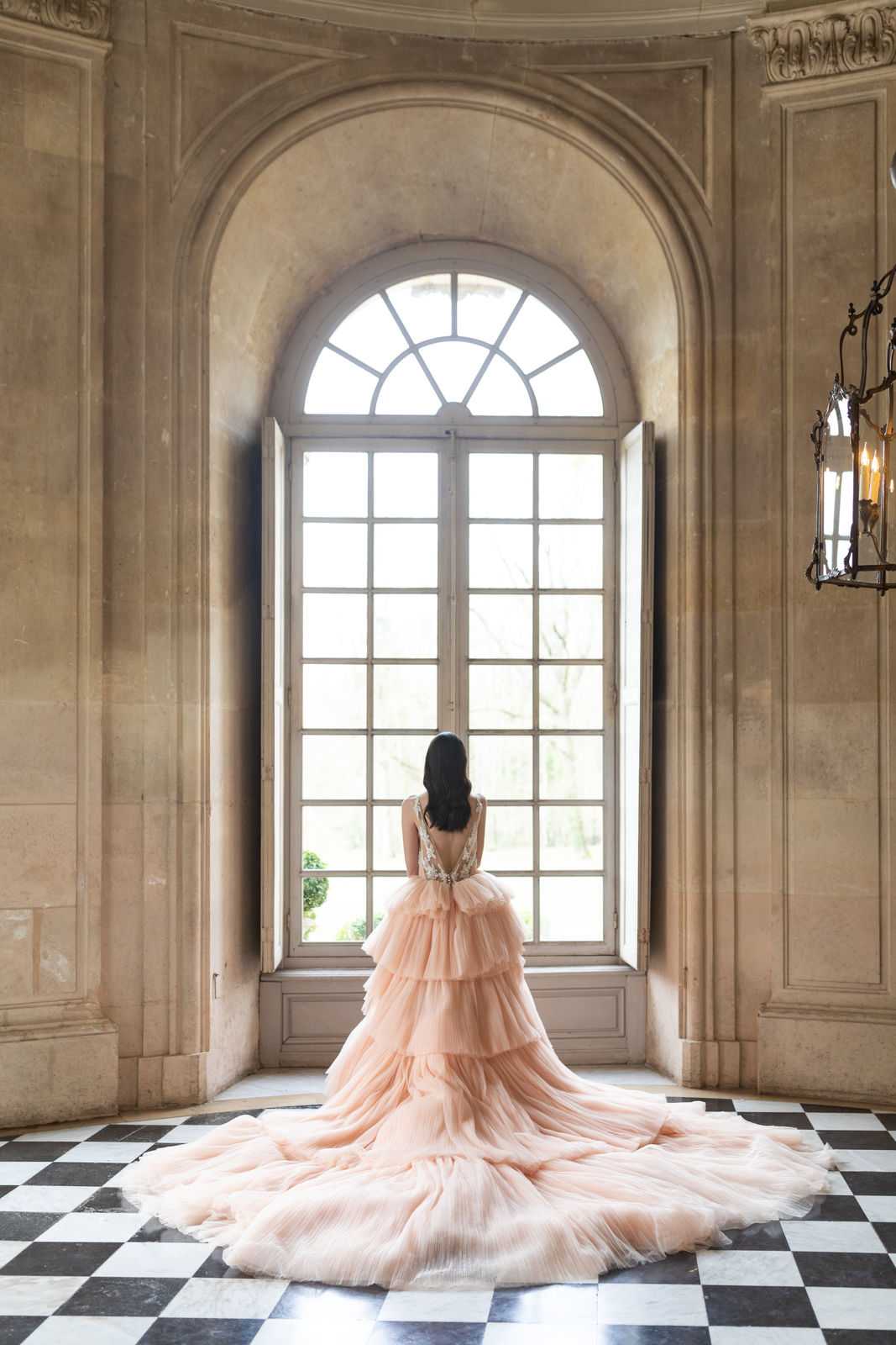 A bridal portrait shot from behind, showing a bride standing at a tall arched window inside a grand French chateau or palace interior. The bride wears a voluminous blush peach tiered tulle ballgown with a deep V-back bodice featuring embellished floral detailing, and the full cathedral-length train spreads across a black-and-white checkered marble floor. Her long dark hair falls loose down her back. The room features ornate carved stone archwork, aged limestone walls, and a wrought-iron candle lantern visible on the right side. The composition is a wide portrait shot centered on the window, emphasizing the dramatic scale of the gown against the architectural setting. Potential venue feature image.