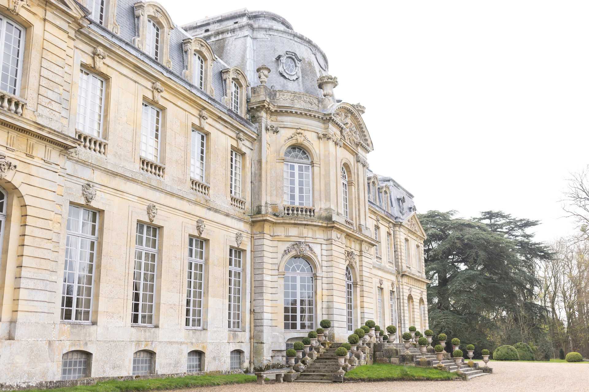 An exterior shot of a French classical chateau facade, shot from a low angle looking along the length of the building. The structure features cream-toned limestone masonry, ornate carved stone detailing, arched windows, balustrades, and a mansard roof with a prominent domed central pavilion. Stone steps lead up from a gravel courtyard to the entrance, flanked by rows of clipped topiary spheres in stone urns. No people are visible in the image. The wide-angle composition emphasizes the architectural scale and symmetry of the building. Potential venue feature image.
