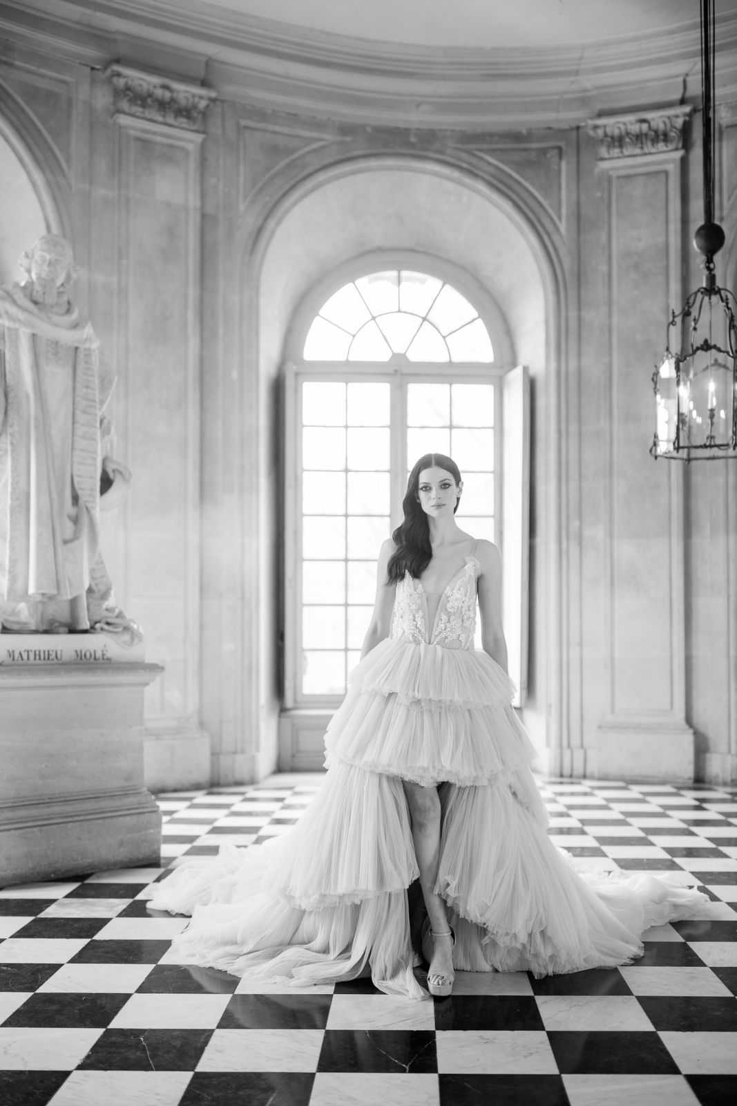 This is a black-and-white bridal portrait shot in a grand classical interior, identifiable by a marble pedestal inscribed 'Mathieu Molé' bearing a sculpted figure, a large arched window with fanlight detail, a wrought-iron lantern chandelier, and a black-and-white checkered marble floor. The bride stands centered before the arched window, wearing a high-low tiered tulle gown with a deep V-neck lace bodice and layered ruffled skirt that trails behind her, paired with strappy heeled sandals. Her dark hair is worn in loose waves over one shoulder, and she faces the camera directly in a composed full-length portrait. The high-contrast B&W tones emphasize the architectural details and the volume of the tiered tulle skirt against the geometric floor pattern. Potential venue feature image.