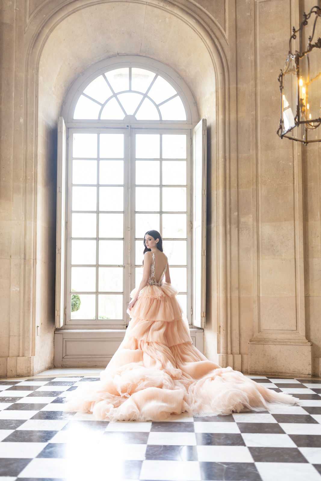 A bridal portrait taken indoors in a grand classical French interior, featuring a bride standing in front of a tall arched French window with natural backlight flooding the room. The bride wears a voluminous blush peach tiered tulle ballgown with an embellished, low-cut open back bodice and an extended cathedral-length train that spreads across the black-and-white checkered marble floor. She is turned slightly, glancing back over her shoulder toward the camera. The room features limestone-colored stone walls with classical molding and pilasters, and a wrought-iron candelabra chandelier is visible in the upper right corner. Full-length portrait shot with the train as a central compositional element. Potential venue feature image.
