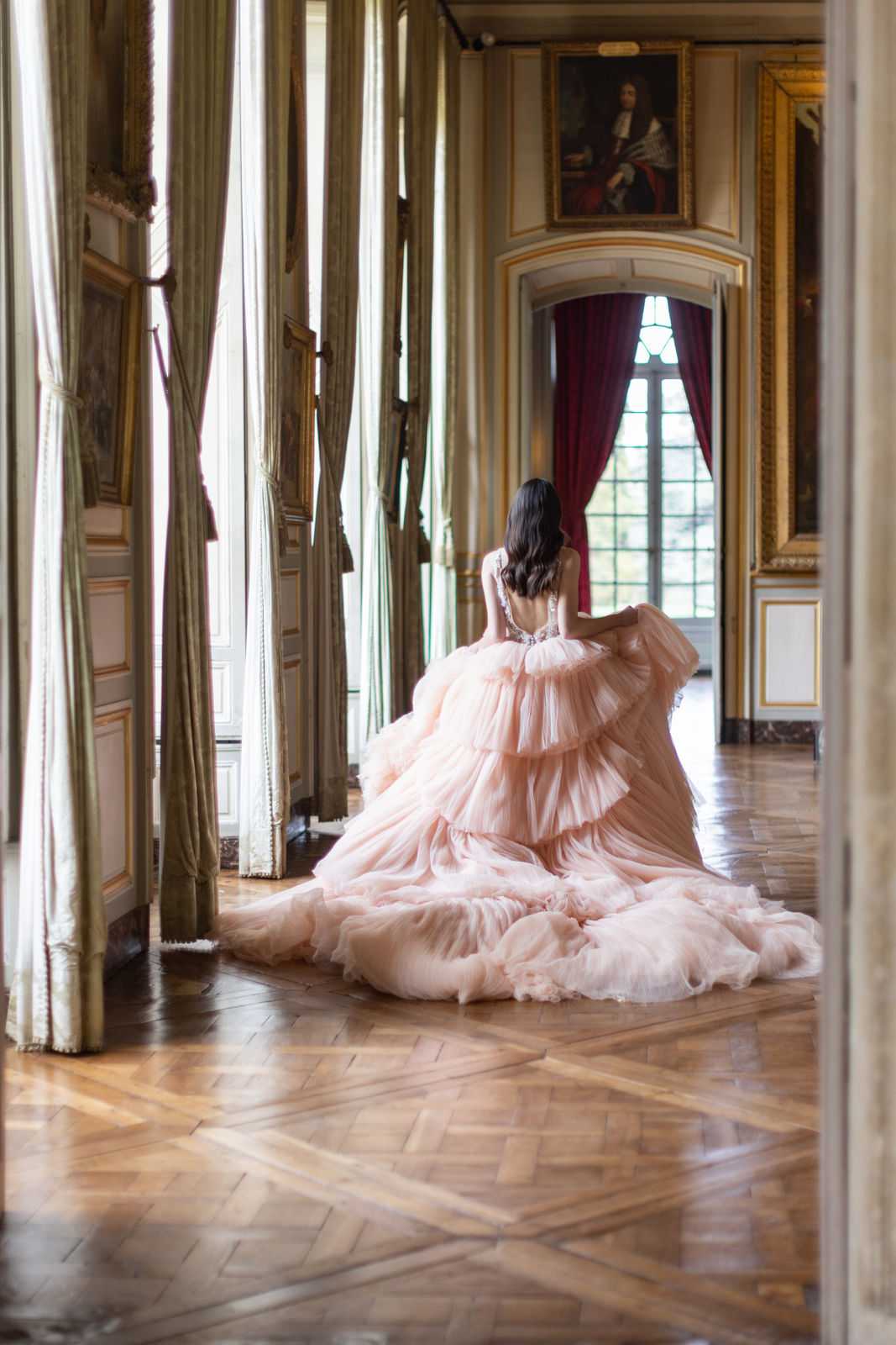 A bridal portrait shot from behind inside a grand palace or chateau gallery, showing a bride walking away from the camera along a long ornate corridor. She wears a voluminous blush pink tiered tulle ball gown with a heavily ruffled skirt and an embellished, open-back bodice, with an extensive train spread across the herringbone parquet floor. The interior features gold-framed oil portraits on the walls, cream and gold molded paneling, floor-to-ceiling windows with sage green curtains and deep crimson velvet drapes, consistent with a classical French royal residence aesthetic. The composition is a wide full-length shot framed through a doorway, with natural window light illuminating the end of the corridor.