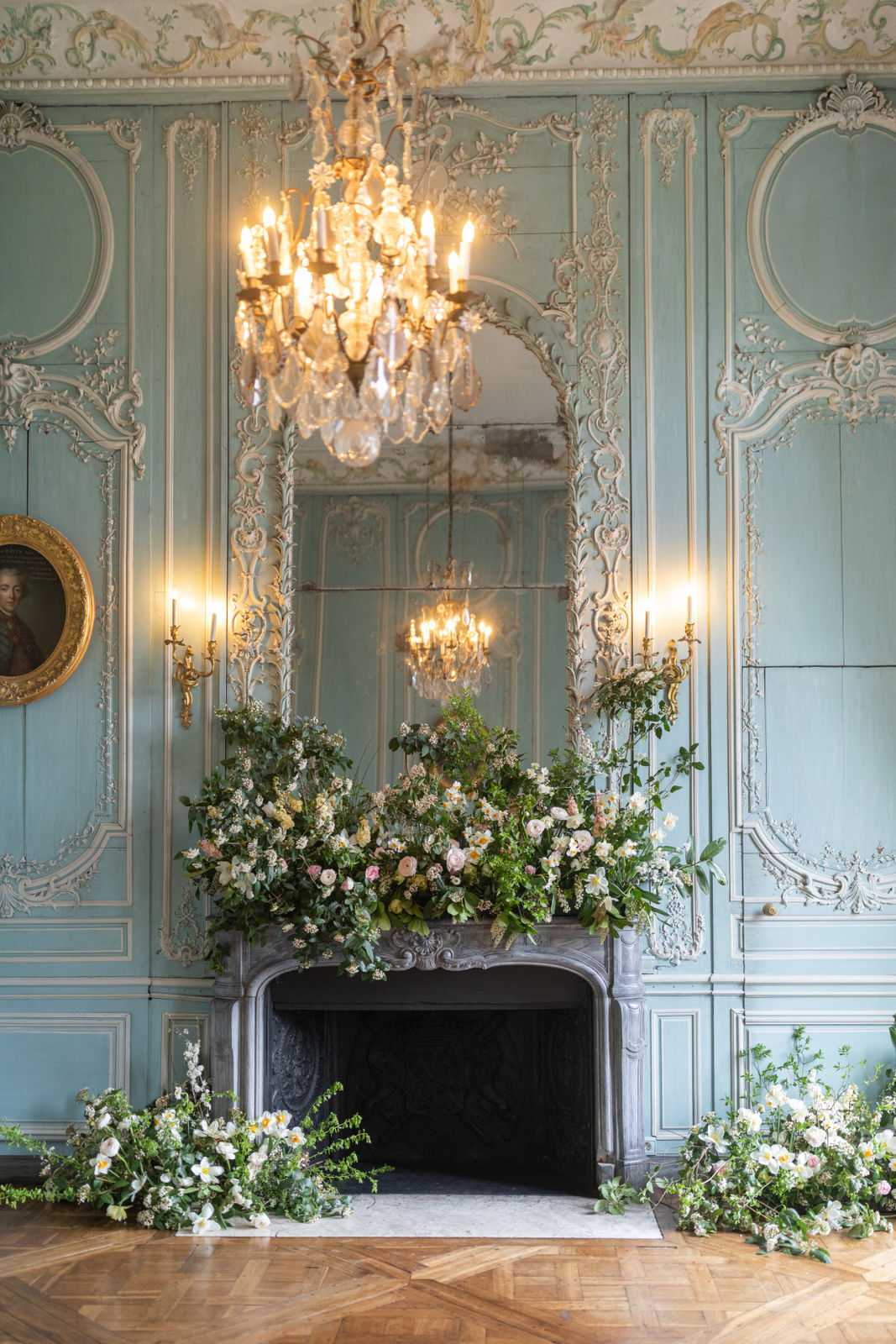 A detailed interior shot of a grand fireplace installation inside a French chateau ballroom, decorated with an abundant floral arrangement in a garden-style, unstructured design. The mantelpiece and surrounding floor are covered in trailing greenery, white anemones, blush and soft pink garden roses, cream ranunculus, and small white blossoms spilling onto the herringbone parquet floor on both sides. The room features powder blue boiserie paneling with white-painted rococo plasterwork, a large ornate mirror above the fireplace, a crystal and gilt chandelier overhead, and twin gold candle wall sconces casting warm light. A small oval portrait painting in a gold frame is visible on the left wall. The composition is a straight-on wide shot framing the fireplace symmetrically within the room. Potential venue feature image.