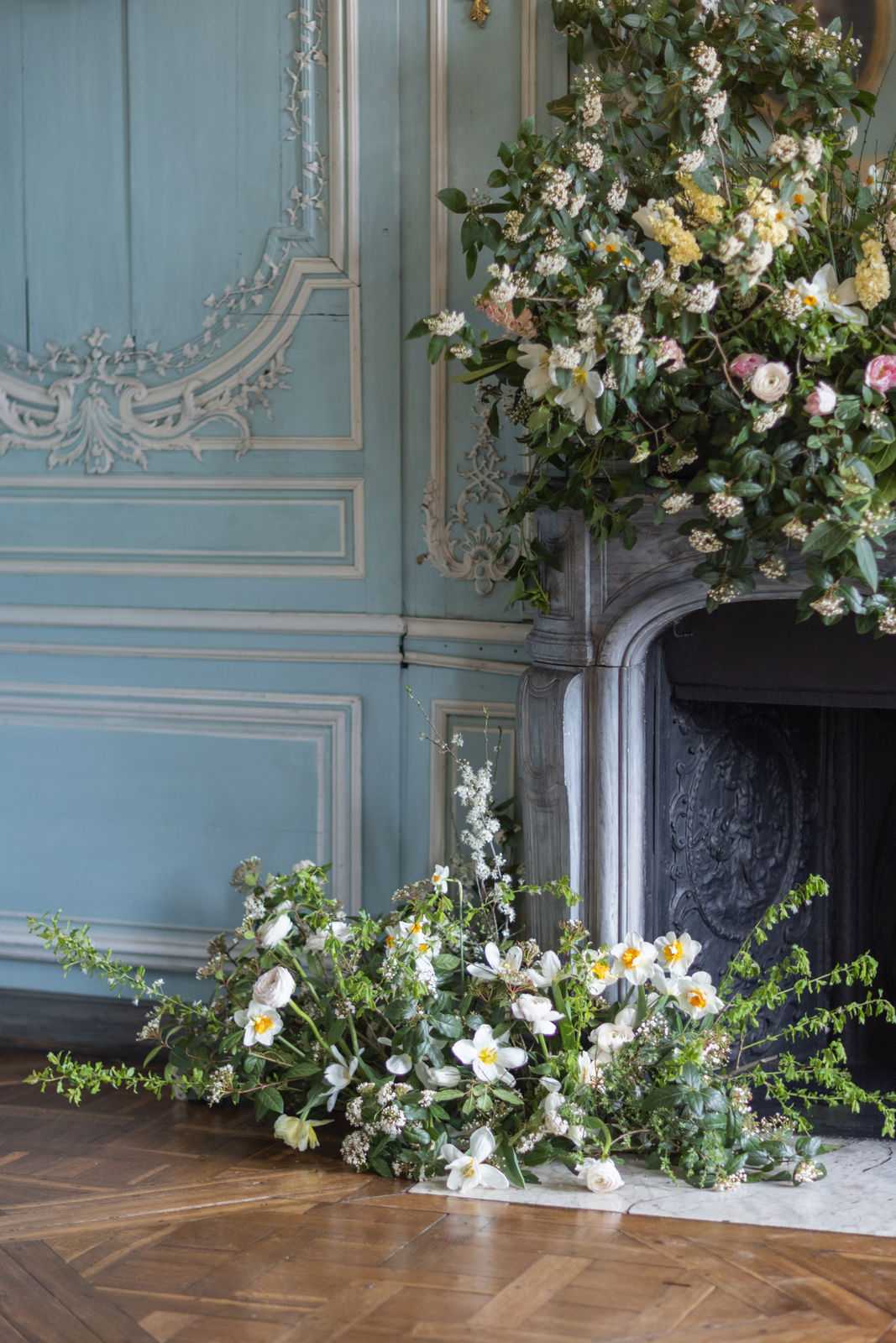 A floral decor detail shot taken indoors in what appears to be a French château or historic ballroom, featuring an ornate grey marble fireplace with an elaborate overflowing floral installation. The arrangement includes white narcissus with yellow centers, blush and pink ranunculus, cream and yellow viburnum-style clusters, white lilies, and loose green trailing foliage and branches, spilling dramatically from the mantel down to the parquet herringbone floor in an intentionally wild, garden-gathered style. The powder blue painted boiserie wall paneling with carved white plaster moldings forms the backdrop. A secondary ground-level floral cluster at the base of the fireplace mirrors the overhead arrangement with white narcissus, small white blooms, and sprawling greenery. The overall floral palette is white, cream, soft yellow, and pale pink against deep green foliage, consistent with a classic French romantic aesthetic. Potential venue feature image.