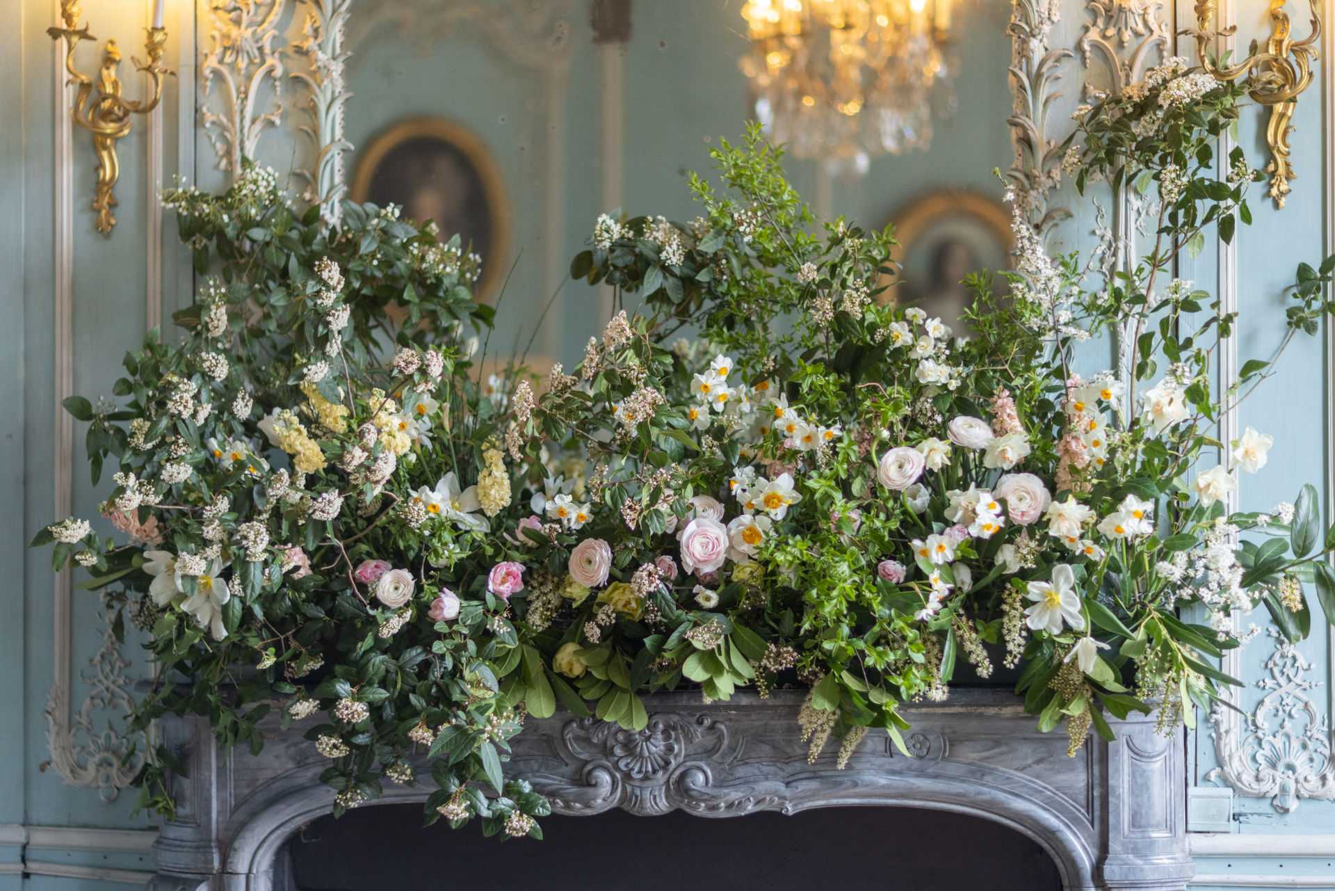 A close-up detail shot of a large floral installation arranged across an ornate carved stone fireplace mantel inside a French chateau-style salon. The arrangement is abundant and garden-style, featuring blush and pale pink ranunculus, white and yellow-centered narcissus, soft pink garden roses, cream stocks, trailing white spirea branches, and dense mixed greenery including leafy vines and small-flowered shrub branches. The overall color palette is soft white, blush pink, cream, and green. The room's decor features duck-egg blue paneled walls with gilded Rococo-style plasterwork, gold wall sconces, and a crystal chandelier visible in the background reflected in a mirror above the mantel. The styling is classic French interior with a loose, garden-gathered floral aesthetic. Potential venue feature image.