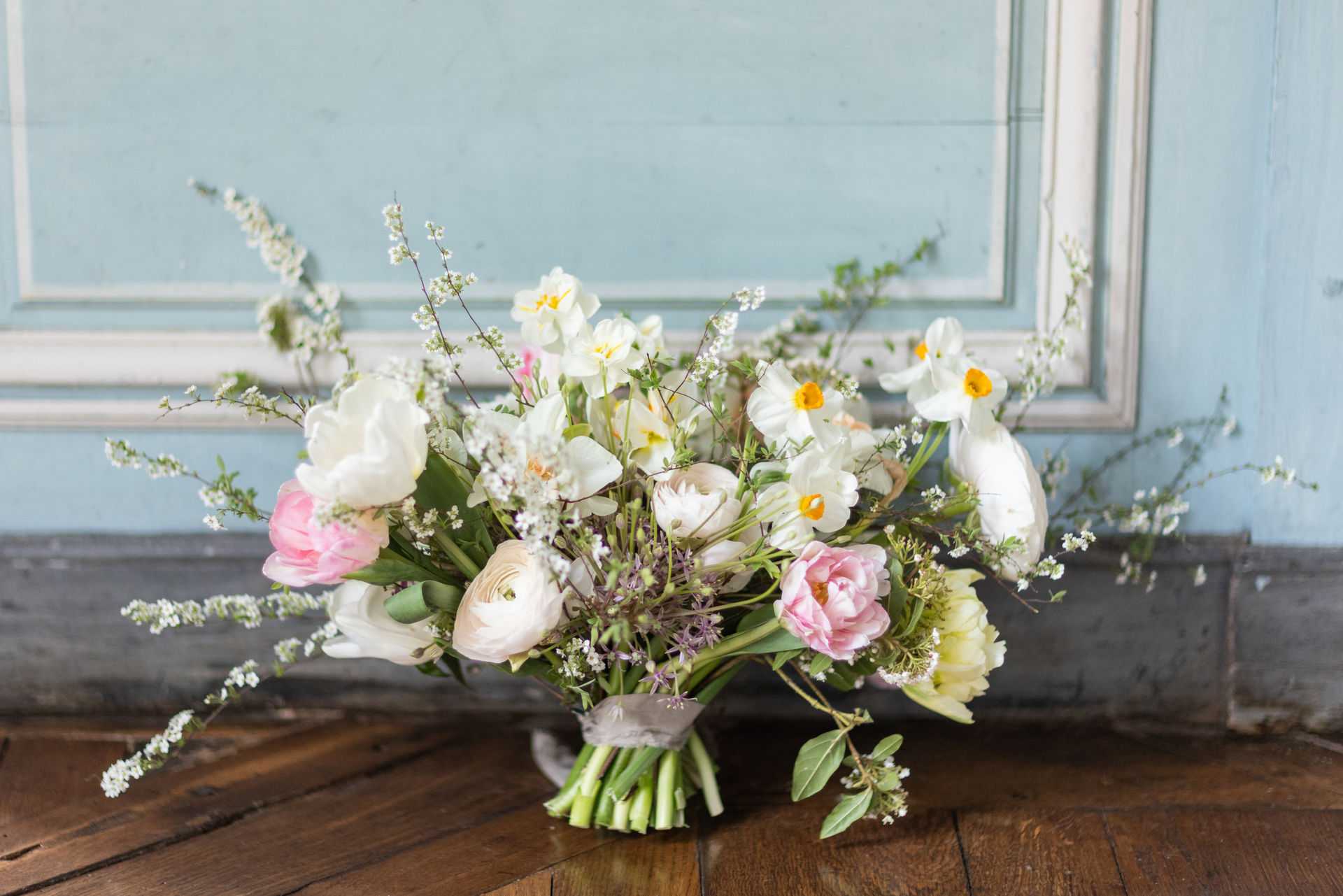 A close-up detail shot of a bridal bouquet resting on a dark wood parquet floor against a pale blue panelled wall interior. The loosely arranged, garden-style bouquet includes white narcissus with yellow centers, pink peonies or tulips, cream ranunculus, white freesias, delicate white sprig blossom, small lavender allium-like filler flowers, and trailing green foliage. The stems are wrapped with a soft grey-silver ribbon. The overall palette is white, blush pink, cream, and soft yellow with green accents, creating a light, airy spring aesthetic.