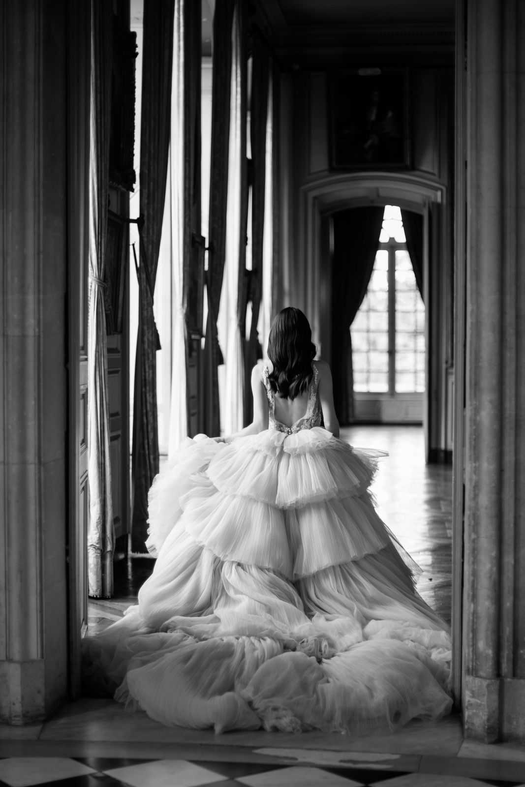 This is a black-and-white bridal portrait taken indoors in what appears to be a grand historic palace or chateau, likely the Palace of Versailles, featuring a long corridor with tall columns, heavy draped curtains, ornate moldings, and large multi-pane windows at the far end providing bright backlighting. The bride is photographed from behind, standing at a doorway threshold, with dark hair worn down in loose waves. She wears a voluminous ball gown with a deeply open back featuring embellished lace or beaded detailing at the bodice, and an extremely full tiered tulle skirt with multiple layered ruffled tiers that pools dramatically across the parquet floor. The image has high contrast with deep blacks in the shadowed corridor and bright whites from the window light, emphasizing the volume of the gown. The composition is a full-length portrait shot framed symmetrically by the doorway columns.
