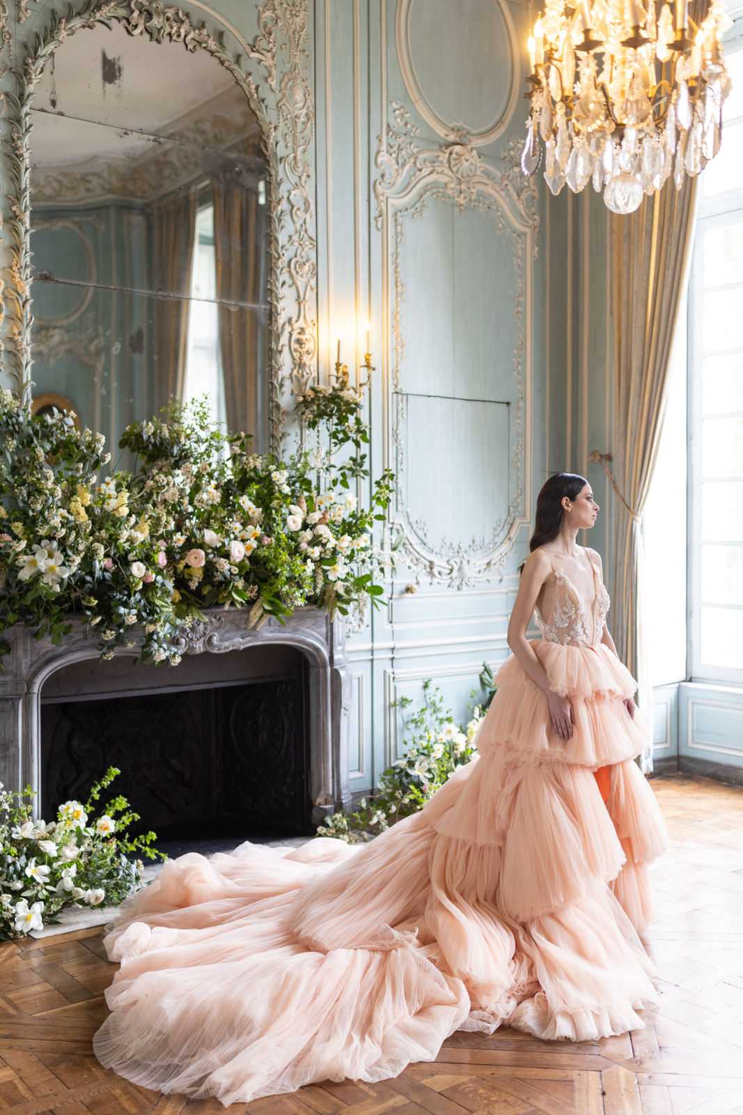 A bridal portrait shot indoors in a French classical interior room featuring powder blue boiserie wall paneling with white-gold rococo plasterwork moldings, a marble fireplace, herringbone parquet flooring, and a crystal chandelier. The bride stands in profile gazing toward a tall window, wearing a blush peach tiered tulle ball gown with an embellished lace bodice and an extended train that fans across the floor. The fireplace mantel and hearth are heavily decorated with an overflowing garden-style floral arrangement of white ranunculus, cream garden roses, yellow blooms, white anemones, and abundant greenery, with additional florals trailing onto the floor. The overall styling palette combines blush, cream, and soft green tones in a classic Parisian interior setting. Potential venue feature image.