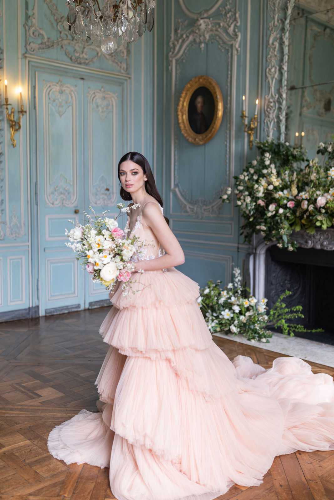 A bridal portrait shoot set inside a French château salon, featuring a bride standing alone on a herringbone parquet floor. She wears a voluminous blush pink tiered tulle ball gown with a long train and an embellished bodice, and holds a loose, garden-style bouquet of white ranunculus, pink peonies, white delphinium, and small yellow blooms with trailing greenery. The room features pale blue-grey boiserie paneling with white carved plasterwork moldings, a crystal chandelier overhead, gilt wall sconces with candles, and an ornate gilt-framed oval portrait painting above a stone fireplace. The fireplace surround is decorated with an oversized lush floral arrangement of white, pink, and cream blooms with abundant greenery. The overall styling theme is classic French rococo with a romantic pastel palette. Medium full-length portrait shot. Potential venue feature image.